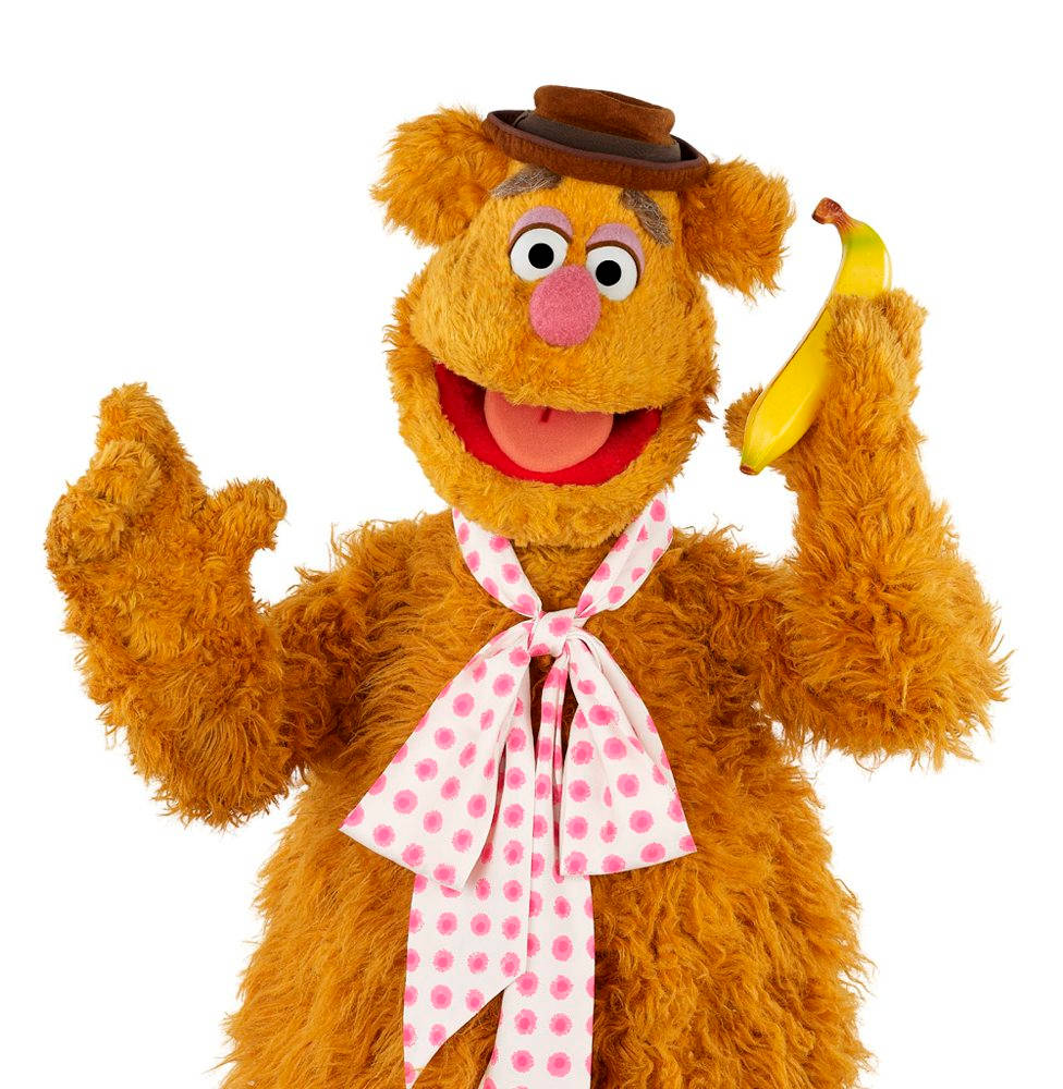 Fozzie Bear 桌布 壁紙 — Fozzie Bear 戴著棕色帽子和粉紅色圓點領結，拿著香蕉，在白色背景前愉快地揮手。