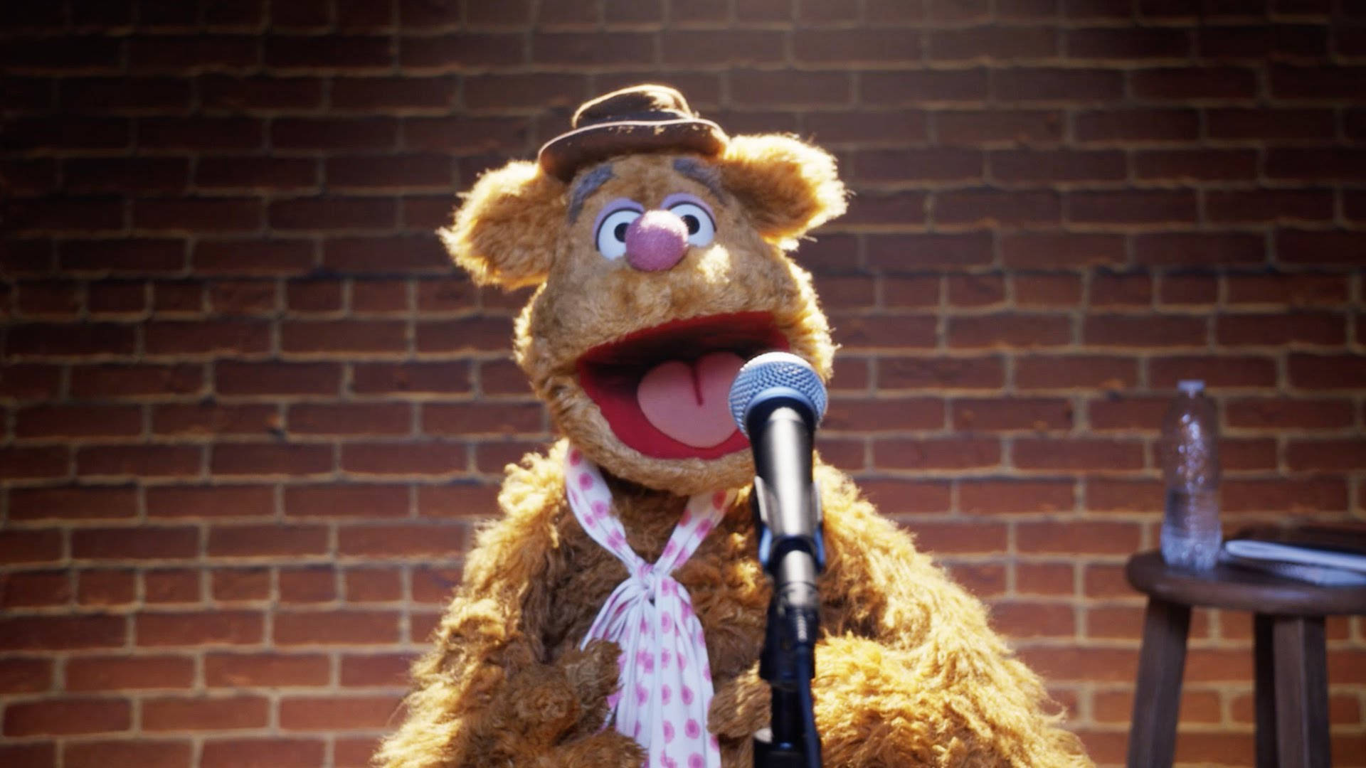 Fozzie Bear 桌布 壁紙 — Fozzie Bear 戴著棕色帽子和圓點圍巾，在磚牆背景下對著麥克風歡快地唱歌。