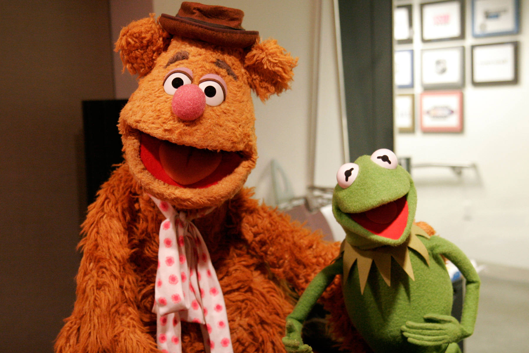 Fozzie Bear 桌布 壁紙 — Fozzie Bear 戴著棕色帽子和圓點圍巾，在溫暖燈光的室內環境中，與Kermit the Frog一起微笑。