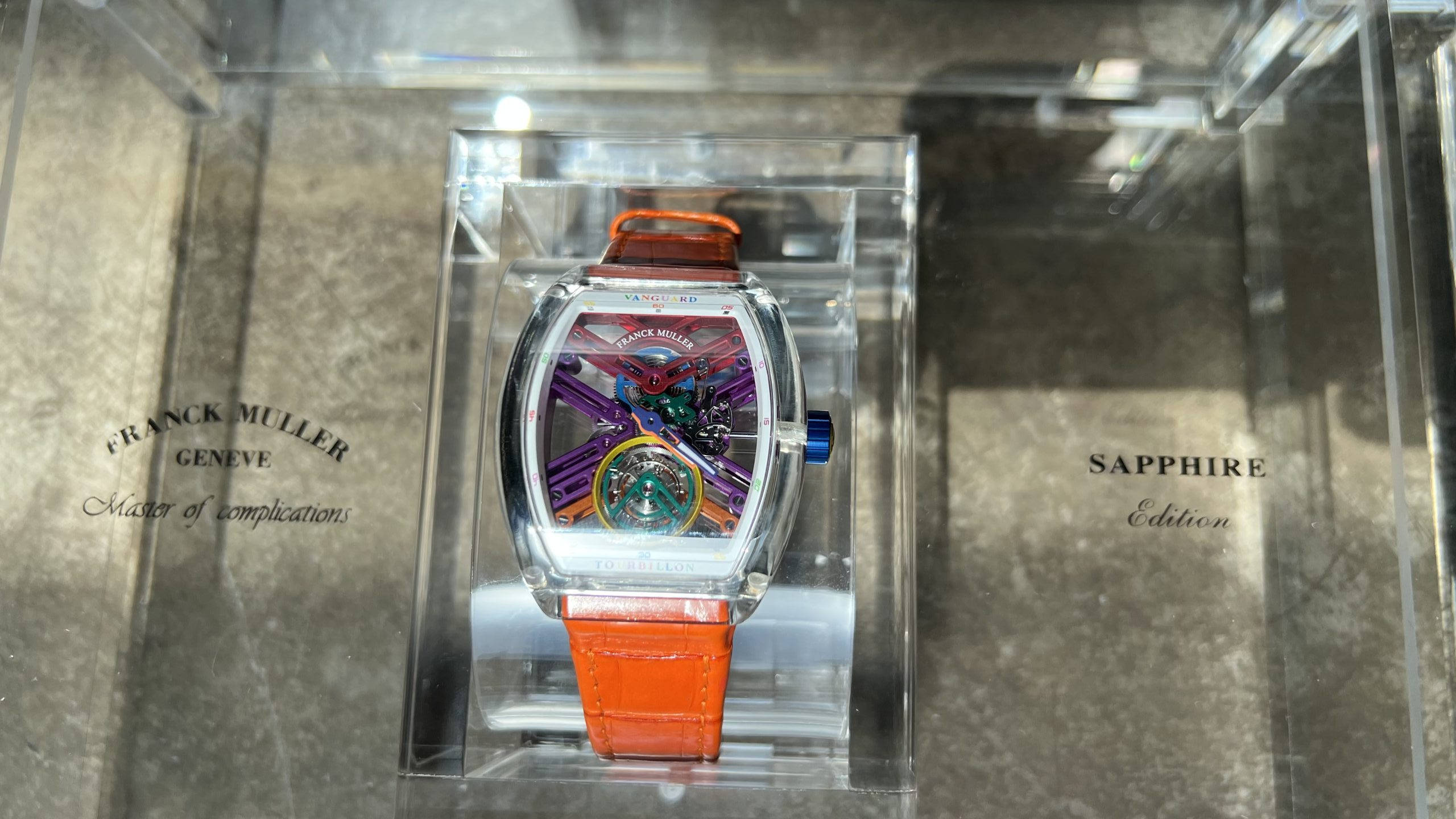 Luxury 桌布 壁紙 — Franck Muller 藍寶石版手錶搭配橙色錶帶，展示在透明錶盒中，置於大理石表面。