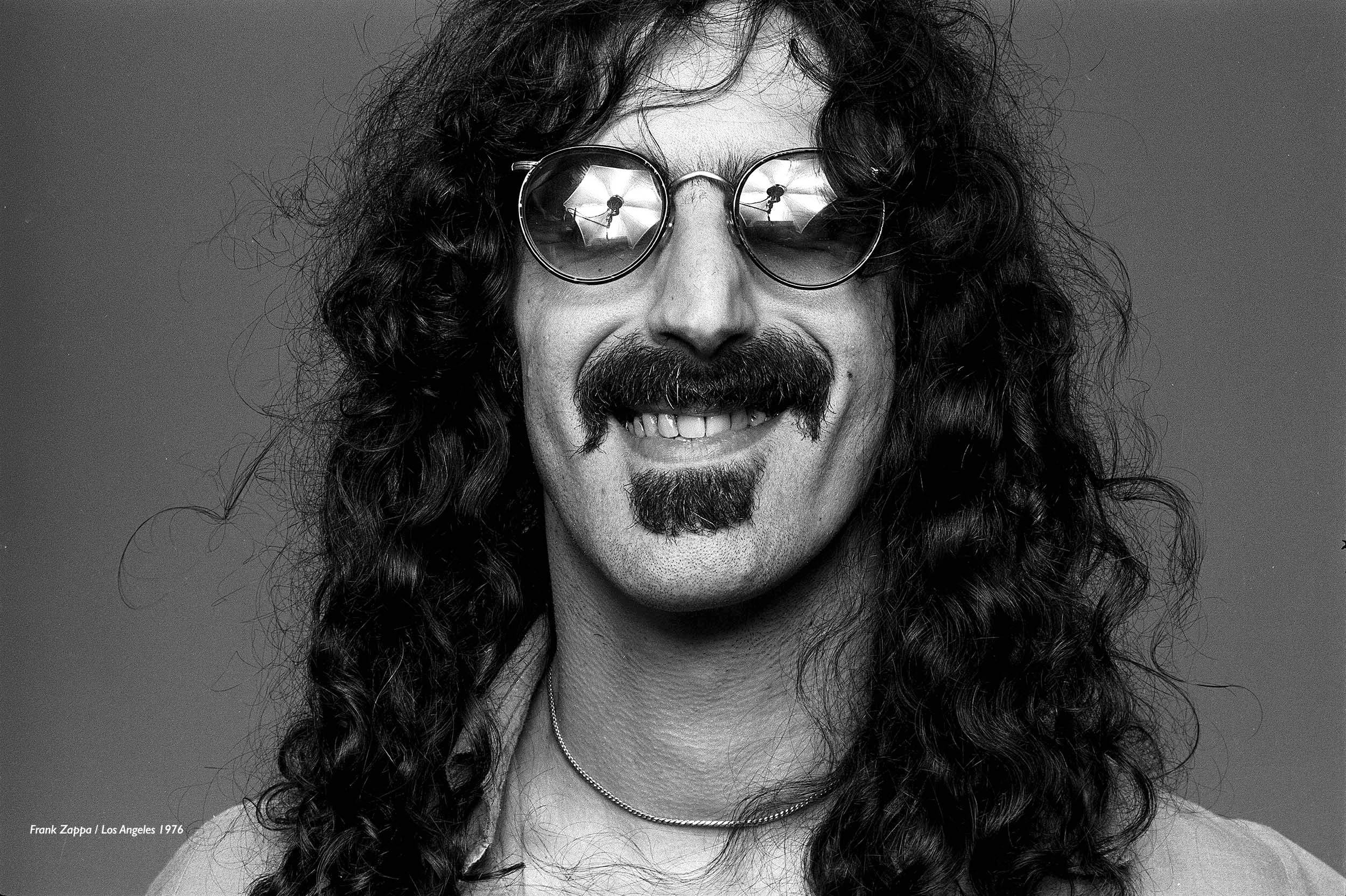 Zappa 桌布 壁紙 — Frank Zappa 戴著圓形太陽眼鏡，鏡片反光，在純灰色背景前微笑。