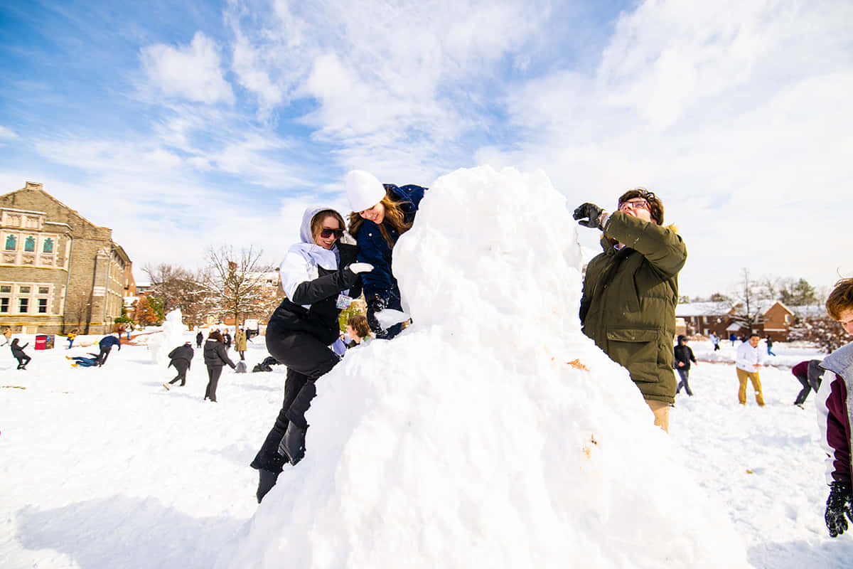  壁紙 — 朋友們在晴朗藍天下，於白雪覆蓋的校園庭院中愉快地建造一個高聳的雪人。