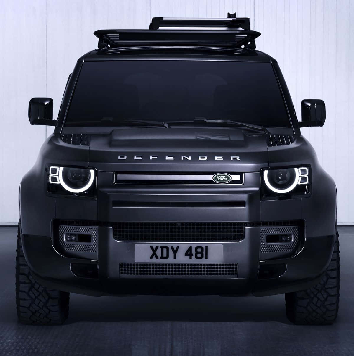 Land Rover Defender 桌布 壁紙 — 白色牆壁前，啞黑色 Land Rover Defender 的正面視圖，配備車頂行李架和發光的圓形頭燈。