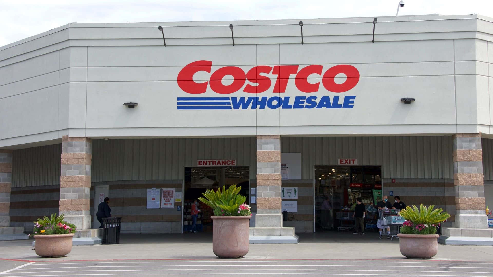 Costco 桌布 壁紙 — Costco Wholesale 商店正面圖，設有紅藍色標誌、入口出口門及盆栽植物，天空多雲。