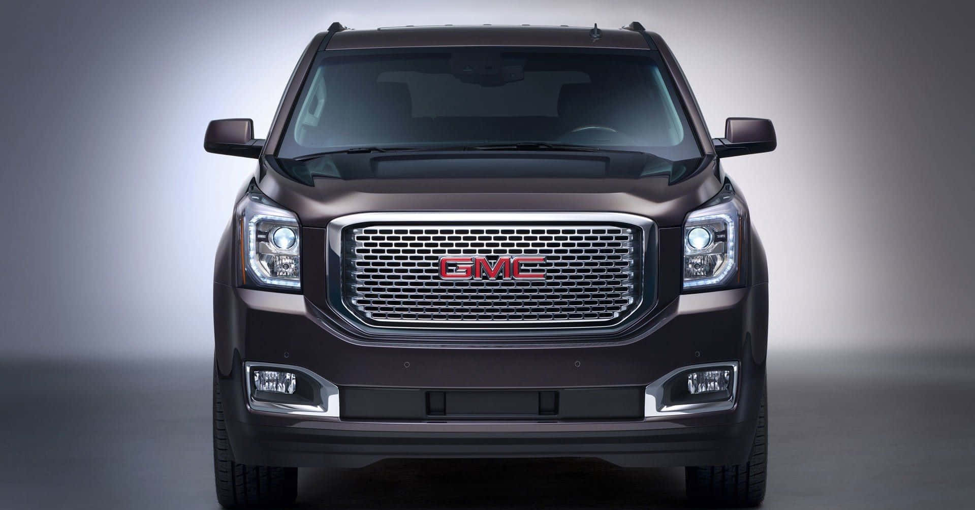 Gmc Yukon 桌布 壁紙 — 深棕色GMC SUV的前視圖，帶有鍍鉻格柵和發光車頭燈，背景為漸變灰色工作室背景。