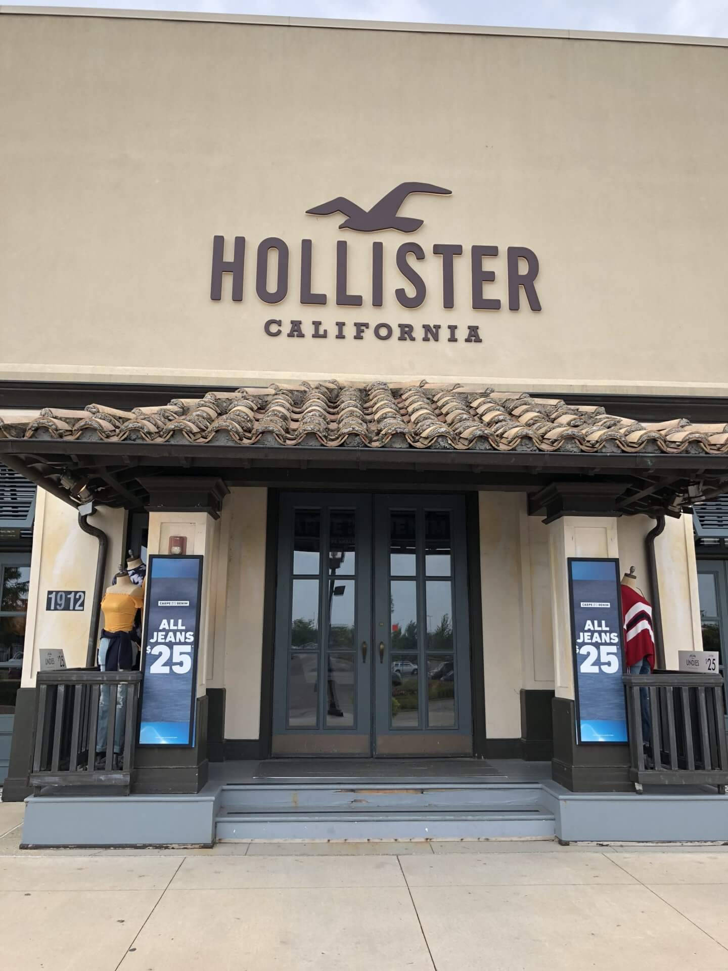 Hollister 桌布 壁紙 — Hollister California 商店的正面視圖，設有瓦片屋頂、人體模型和所有牛仔褲 25 減價標誌。