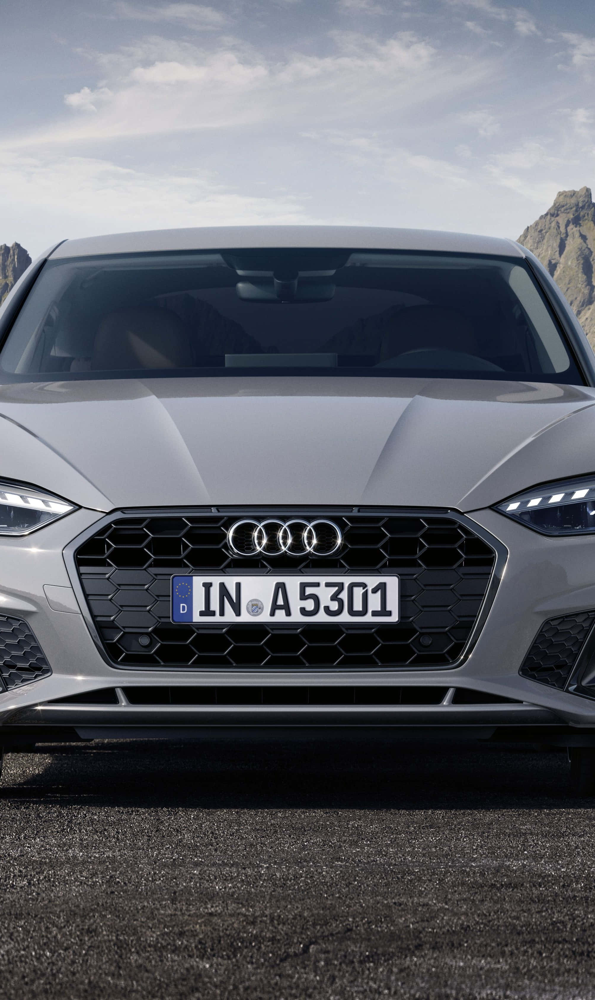 Audi A5 桌布 壁紙 — 銀色奧迪汽車的前視圖，停在瀝青路上，背景有岩石山脈和多雲的天空。