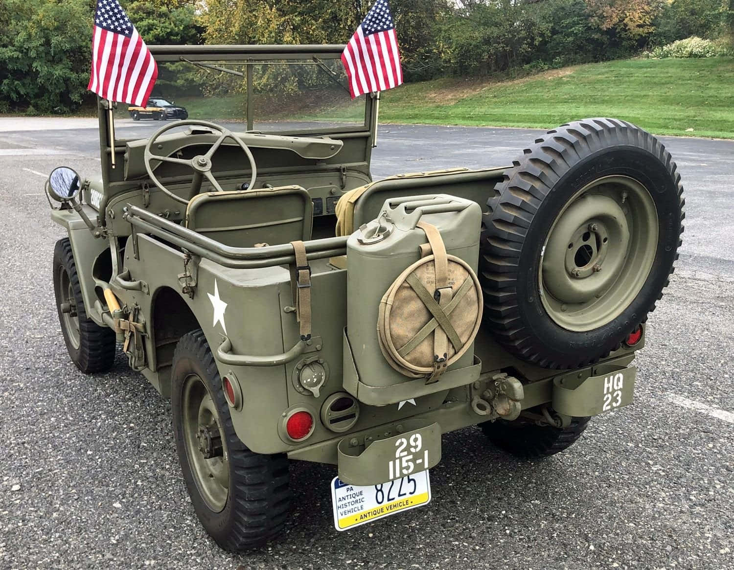 Jeep Willys 桌布 壁紙 — 復古橄欖綠軍用吉普車，帶有美國國旗和備用輪胎，停在靠近草地樹木的瀝青路上。