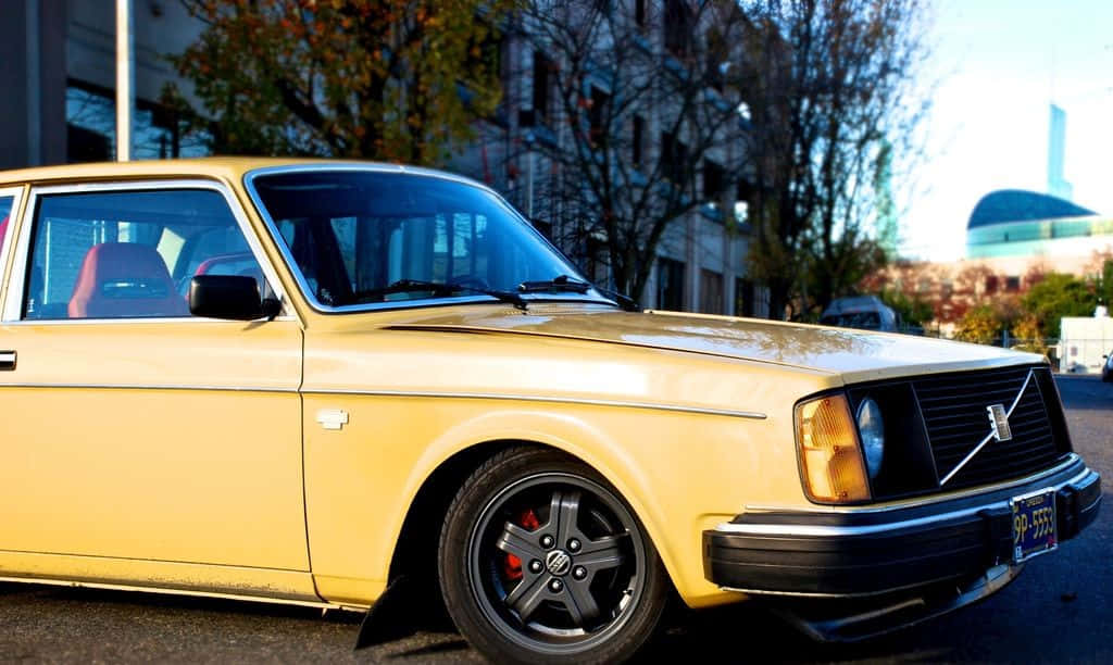 Volvo Pv544 桌布 壁紙 — 一輛復古黃色富豪轎車，配備黑色輪圈和紅色制動卡鉗，在明亮的陽光下停泊在城市街道上。
