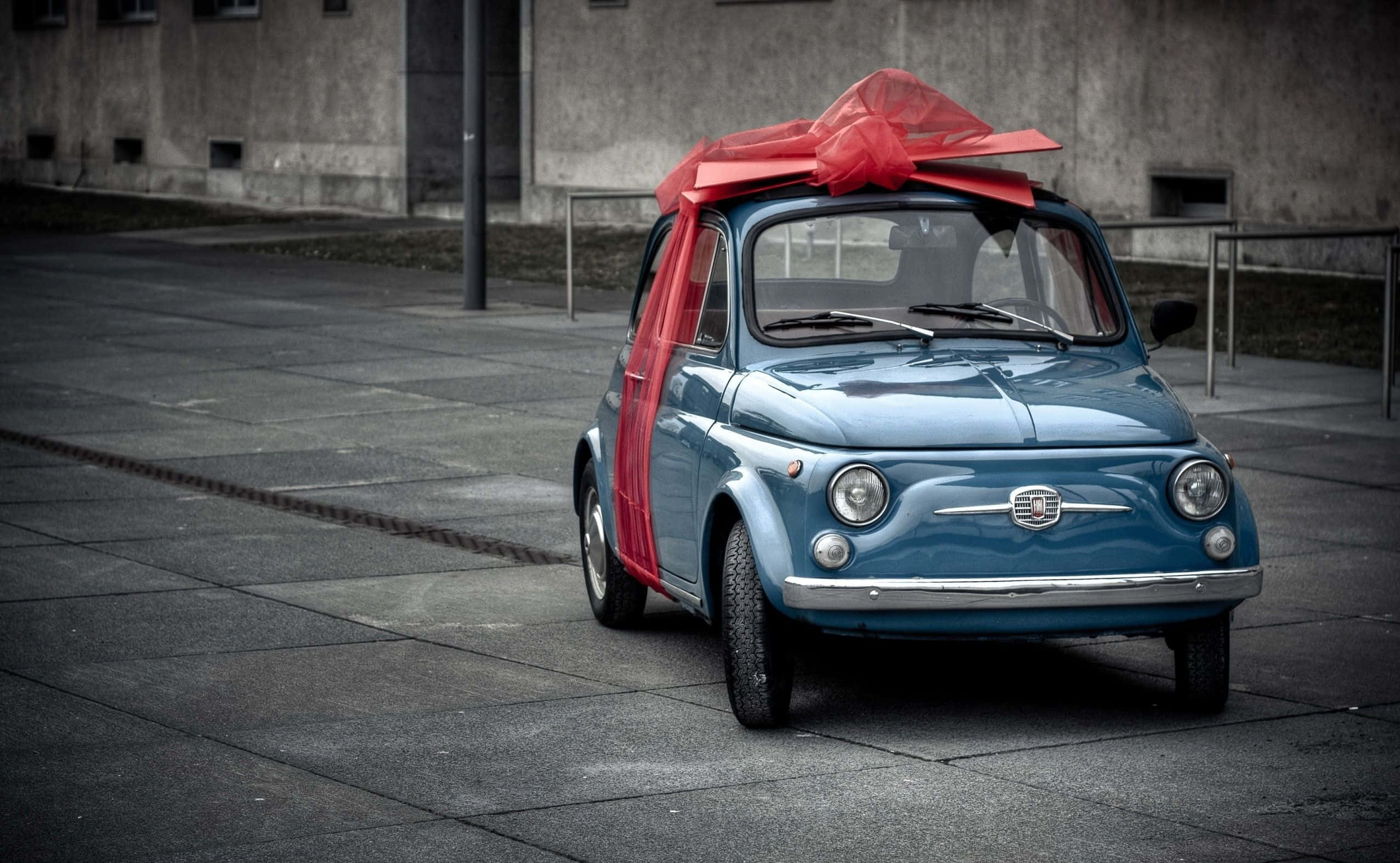 Fiat 500l 桌布 壁紙 — 一輛復古藍色飛雅特500，飾有一個大紅色蝴蝶結，停在灰色混凝土廣場上，背景是風化的工業建築。