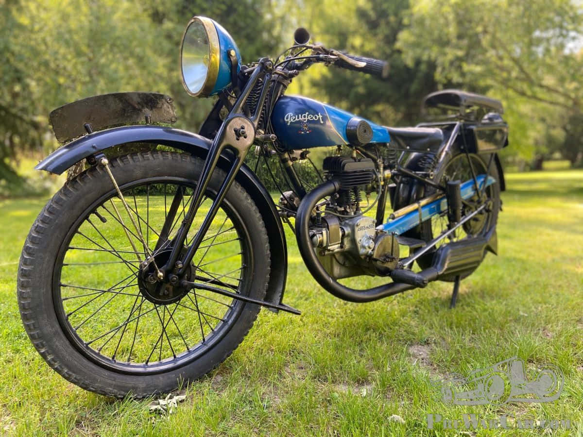 Peugeot Motorcycle 桌布 壁紙 — 復古藍色Peugeot摩托車帶有鍍鉻裝飾，停放在陽光照射的綠色草地上，周圍樹木環繞。