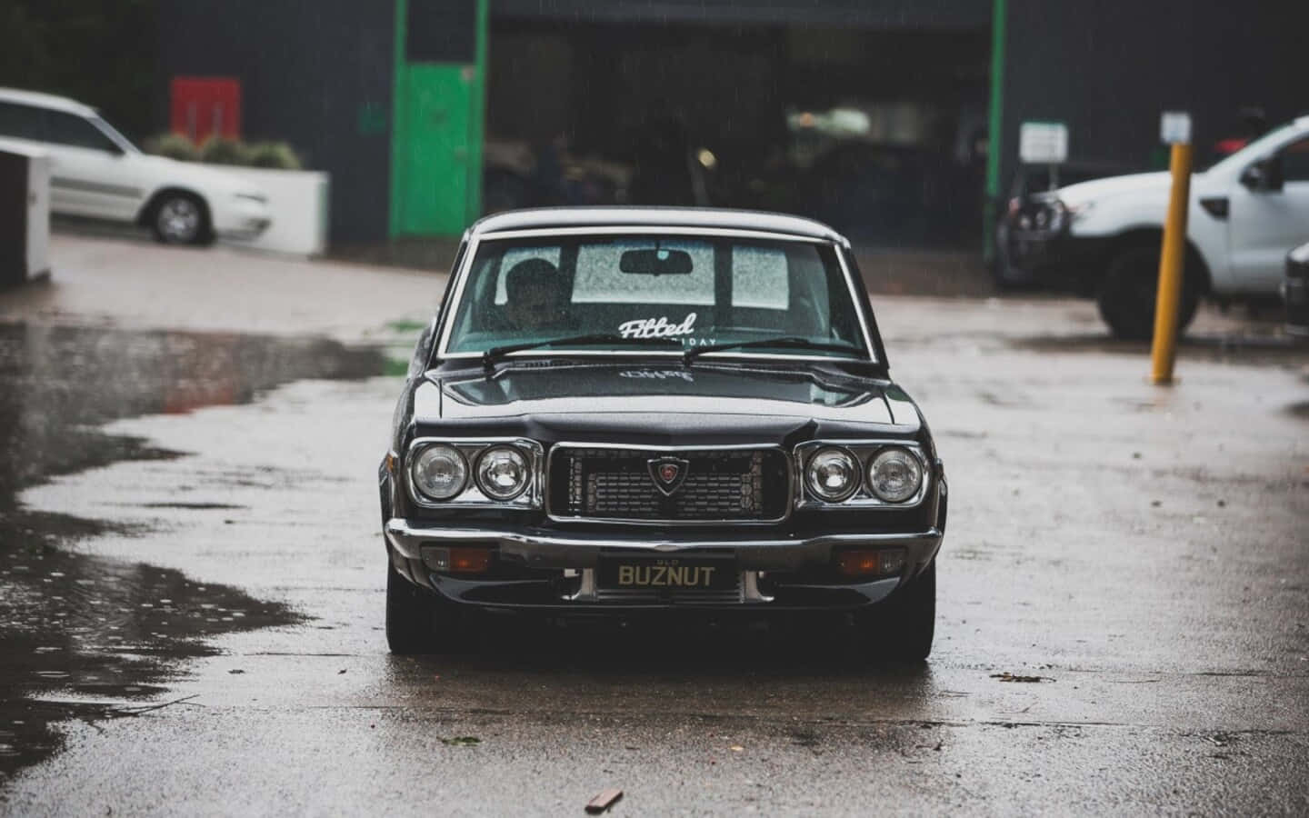 Mazda Rx-3 桌布 壁紙 — 一輛帶有「Fitted」貼紙的復古黑色汽車，在陰天停放在濕漉漉的瀝青場地中央。