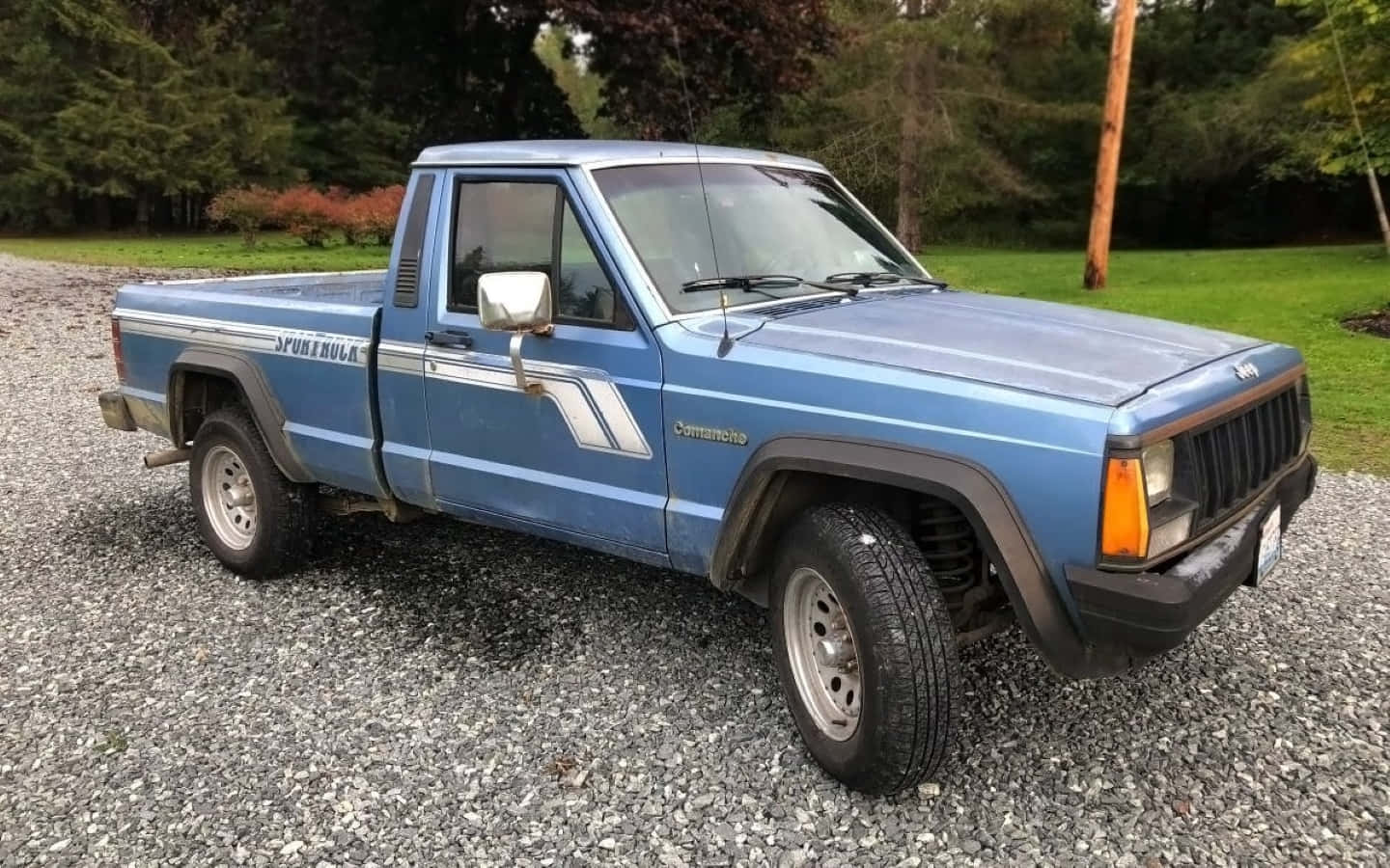 Jeep Comanche 桌布 壁紙 — 一輛復古藍色Jeep Comanche皮卡車停在碎石車道上，周圍環繞著綠樹和草地。