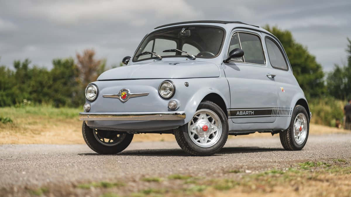 Fiat 500 桌布 壁紙 — 一輛復古淺藍色Fiat 500 Abarth停在碎石路上，背景有綠樹，天空多雲。