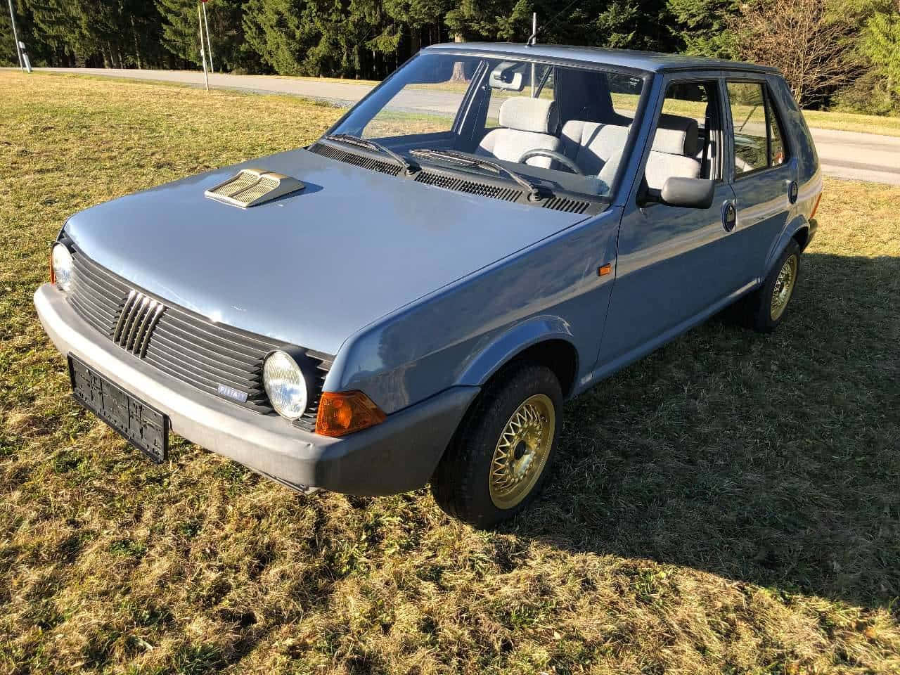 Fiat Ritmo 桌布 壁紙 — 一輛復古藍色飛雅特烏諾停放在乾草地上，背景有樹木，在明亮的陽光下。