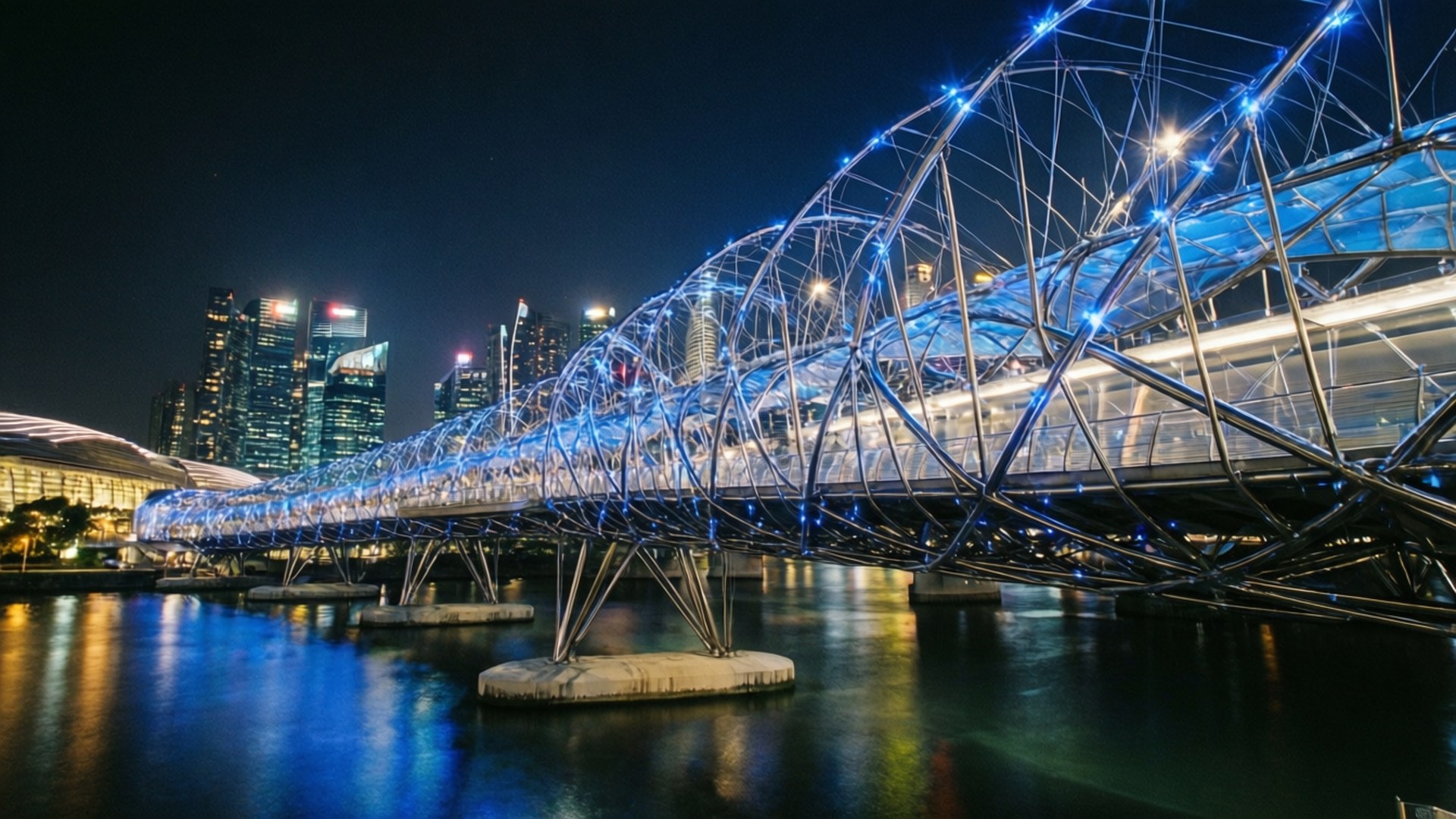 Helix Bridge Night 壁紙 — 一條未來主義的藍色照明行人天橋在平靜的水面上拱起，背景是夜晚發光的摩天大樓。
