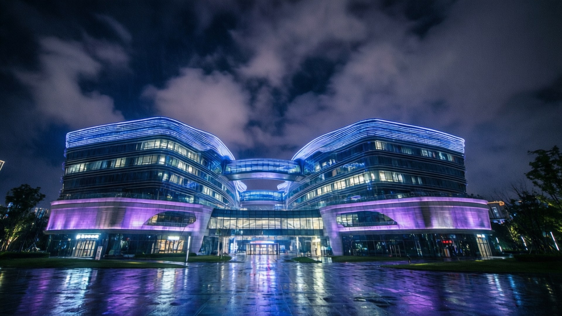 Science City Night Lights 壁紙 — 一座未來主義的建築群在陰雲密布的夜空中閃爍著藍色和紫色的光芒，其倒影在濕漉漉的人行道上閃爍。