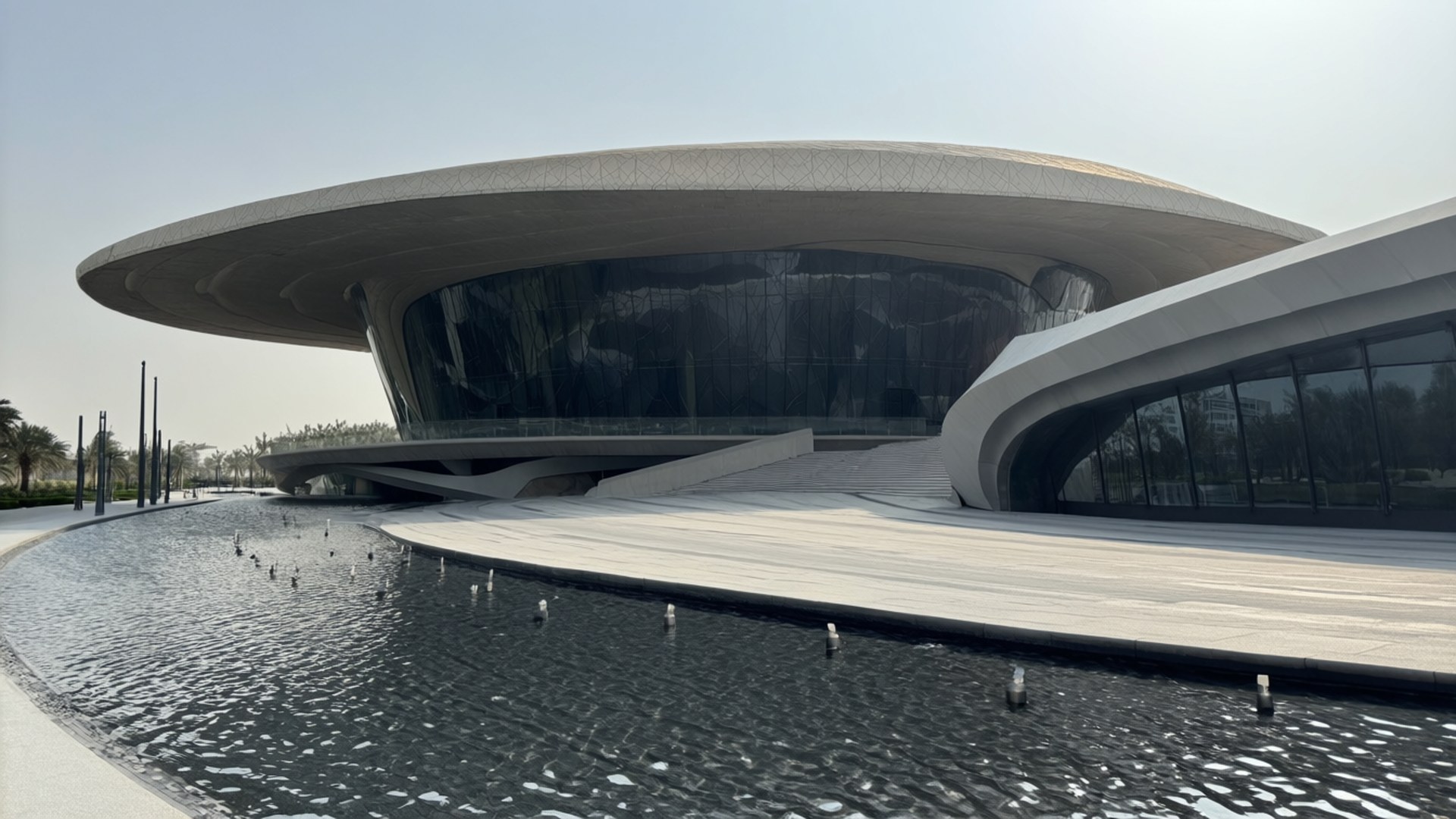 Al-Salem Cultural Centre 壁紙 — 一座未來主義的弧形建築，玻璃牆和白色屋頂，坐落在清澈天空下的反射水景旁。