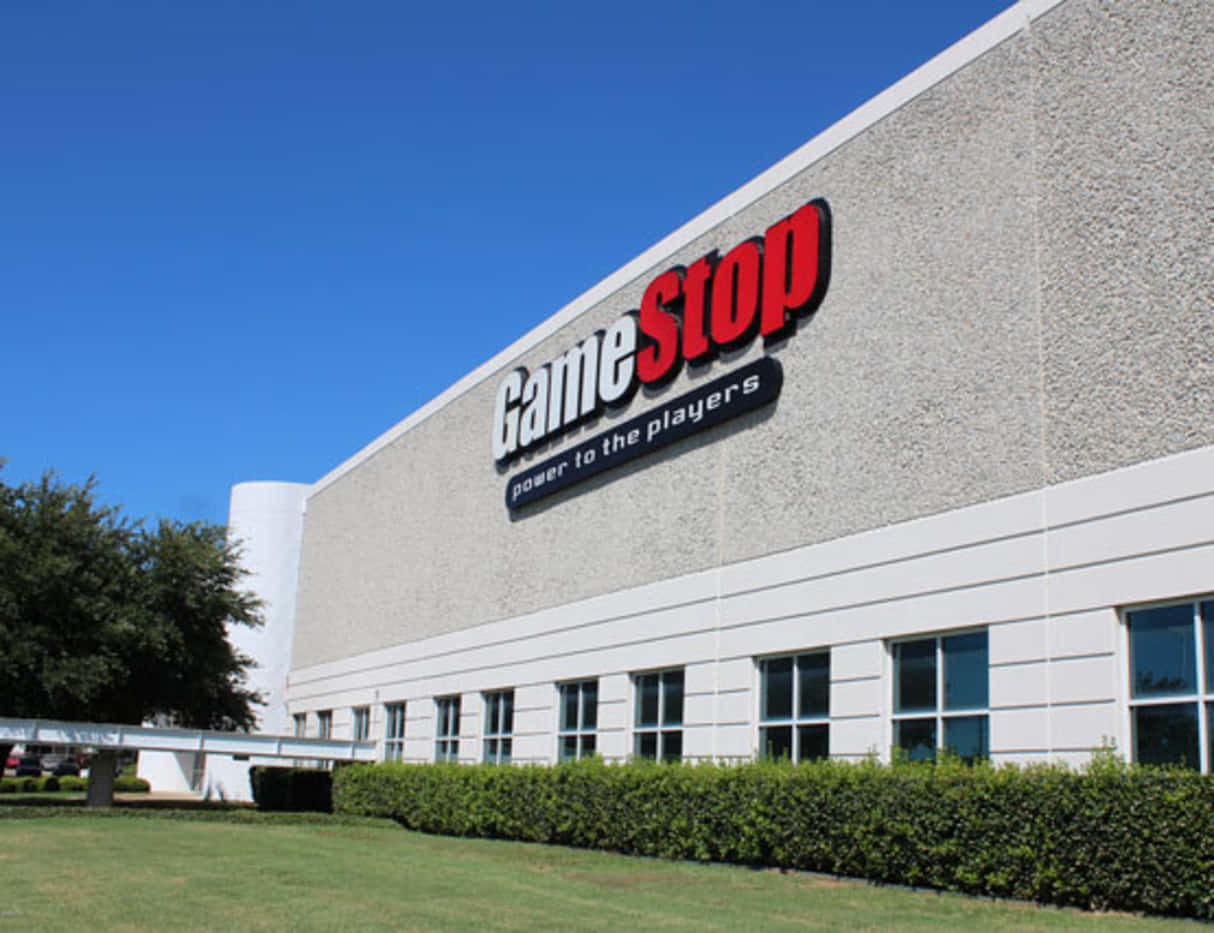 GameStop 桌布 壁紙 — GameStop 商店建築物，在晴朗藍天下，前景有綠色籬笆和樹木。