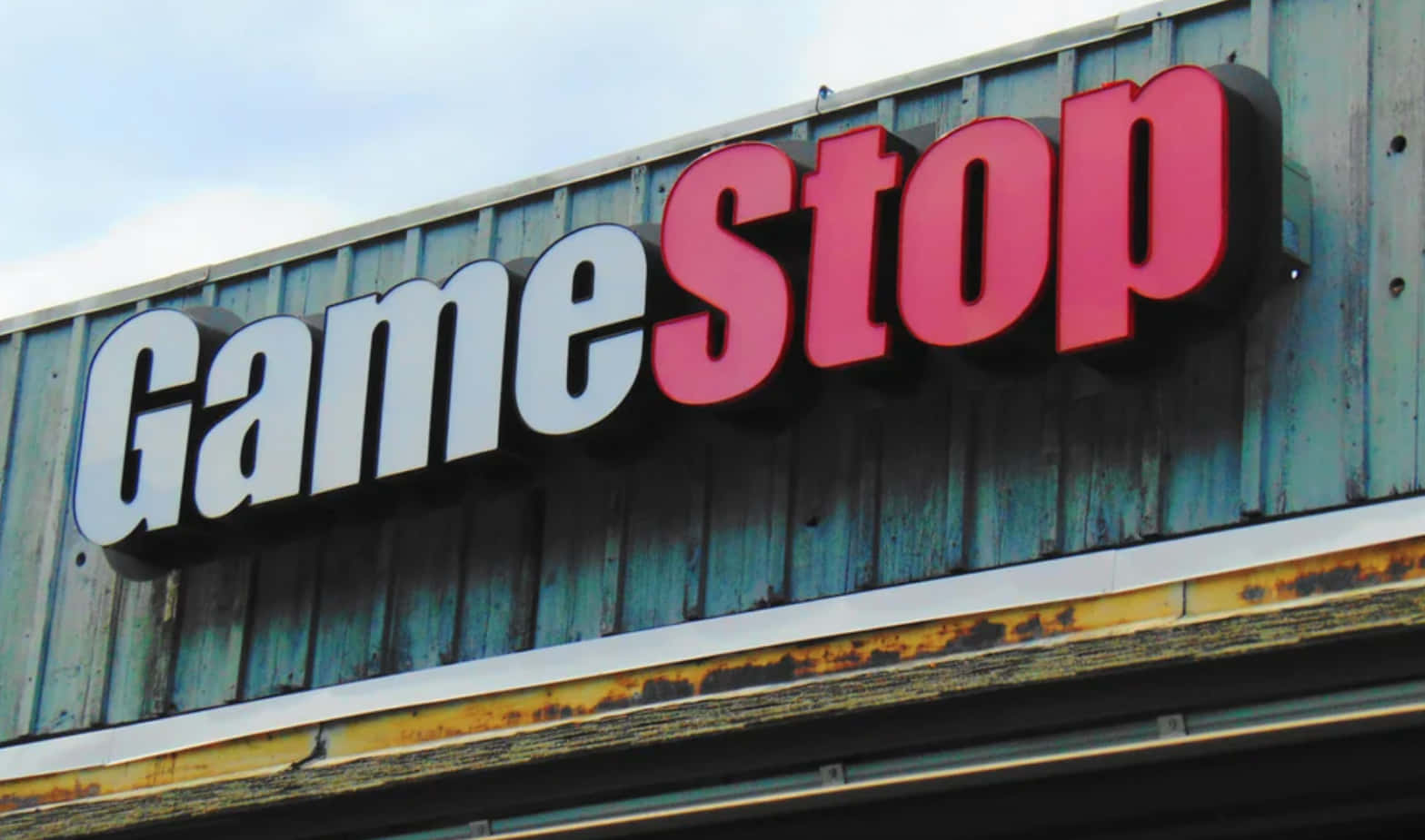 GameStop 桌布 壁紙 — GameStop 商店招牌，白色和紅色字母安裝在風化的藍色金屬壁板上，天空多雲。