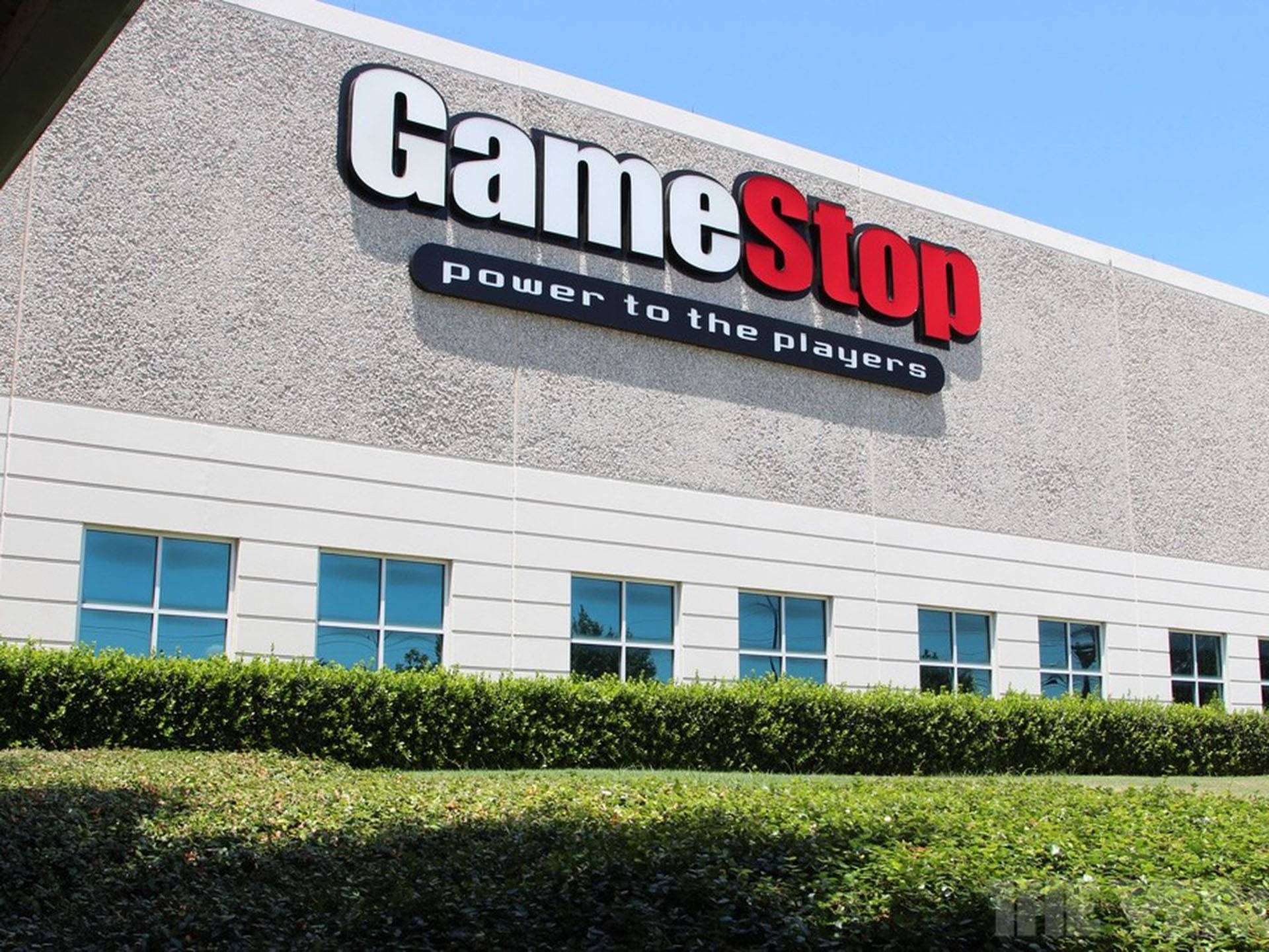 GameStop 桌布 壁紙 — GameStop 店鋪外牆，在晴朗藍天和修剪整齊的綠色籬笆襯托下，設有醒目的紅白招牌。