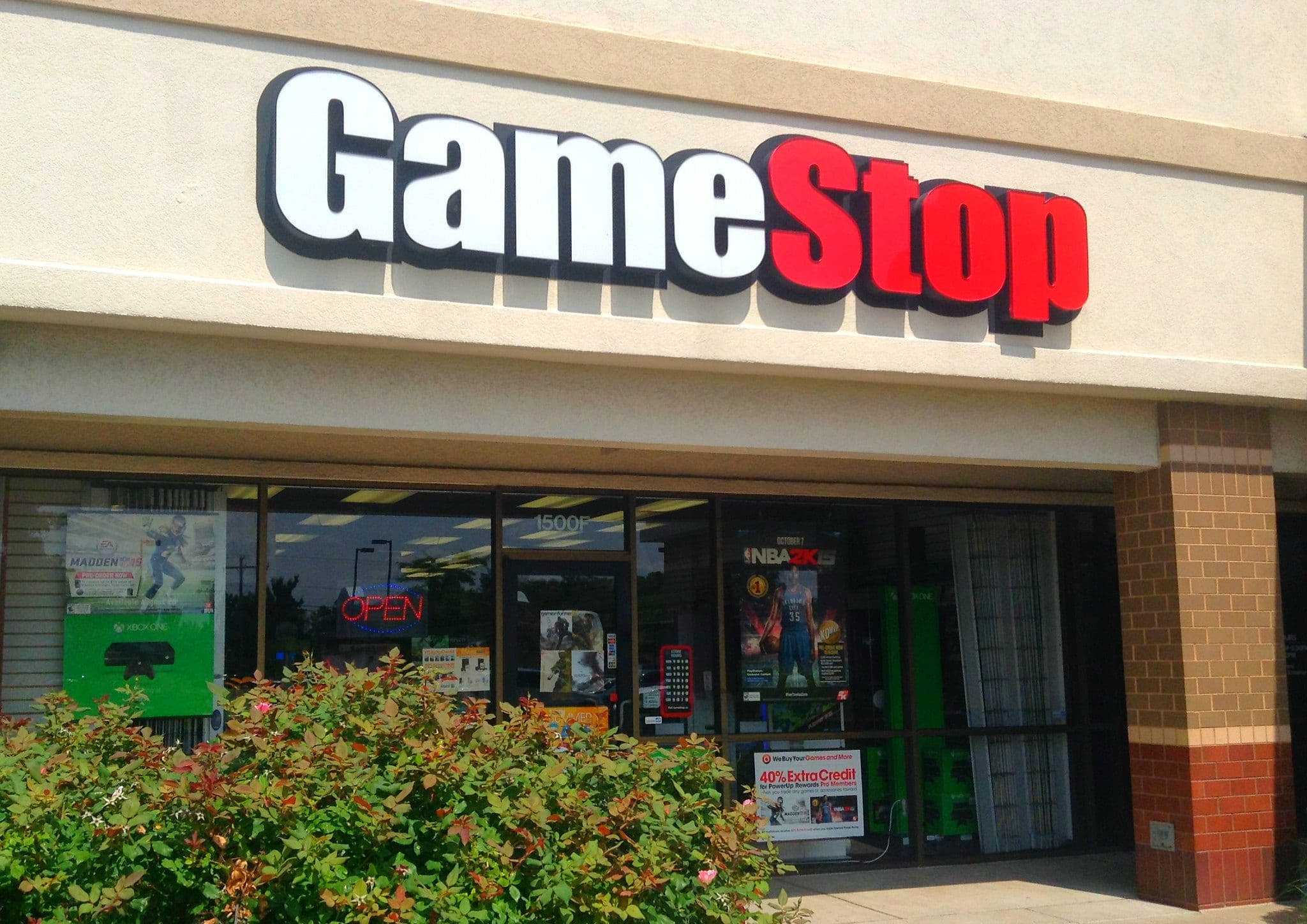 GameStop 桌布 壁紙 — GameStop 店鋪正面，帶有醒目的紅色和白色標牌，玻璃窗展示遊戲海報和營業標誌，周圍環繞著綠色灌木，在明亮的日光下。