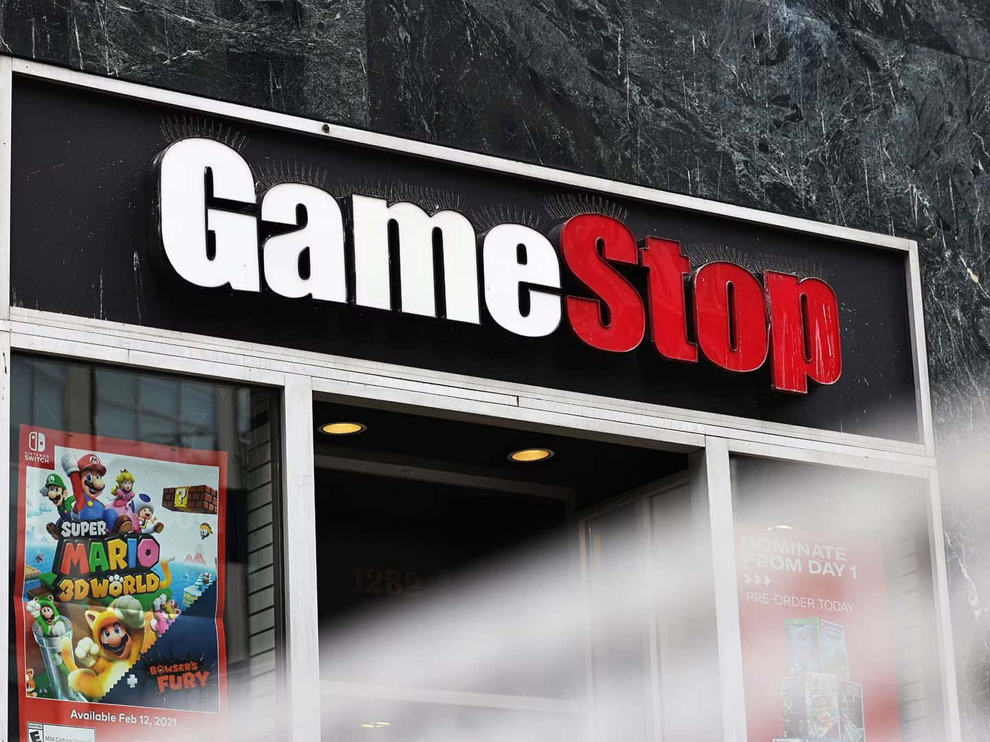 GameStop 桌布 壁紙 — GameStop 店面，採用大膽的紅白招牌，櫥窗內展示 Super Mario 3D World 海報，背景為深色大理石外牆。