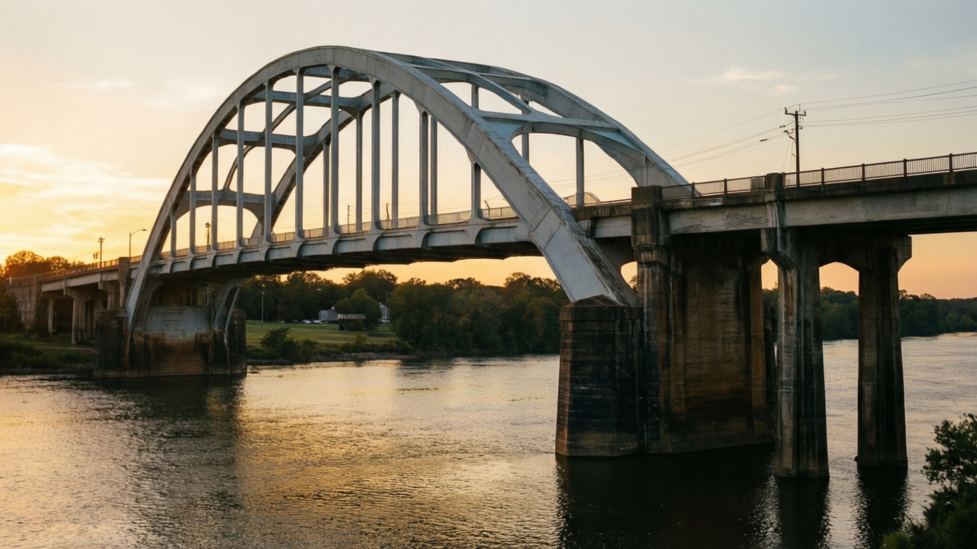 Edmund Pettus Bridge 壁紙 — 一座鋼拱橋橫跨平靜的河流，金色夕陽下溫暖的光線反射在水面上，兩岸樹木成行。