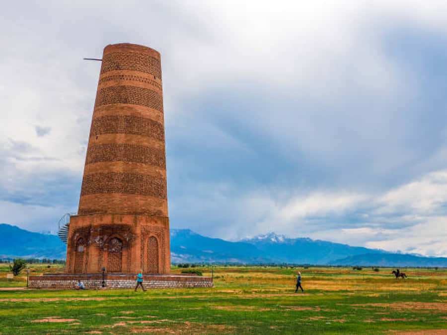 Kyrgyzstan 桌布 壁紙 — 一座高大的古老磚塔孤獨地矗立在廣闊的綠色田野中，遠方有白雪覆蓋的山脈，天空多雲。