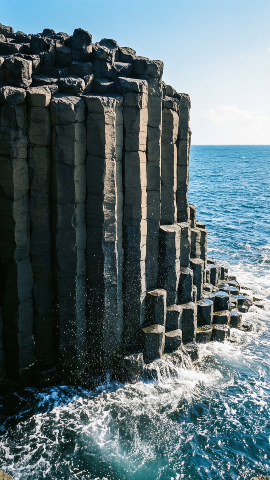 Jusangjeolli Columns 壁紙 — 高聳的六角形玄武岩柱從碧綠色的海水中升起，在清澈的藍天下，海浪拍打著它們崎嶇的底部。
