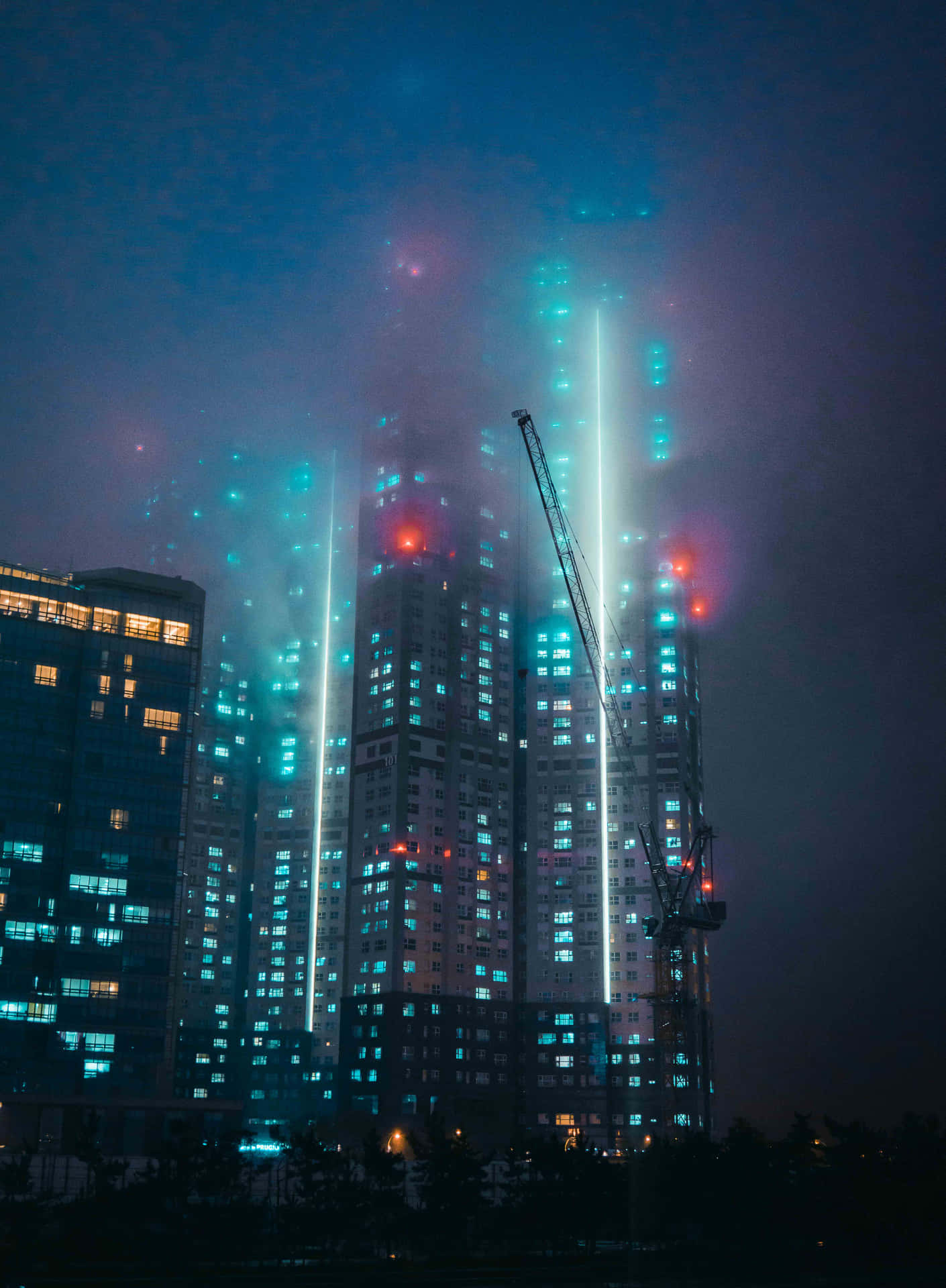 壁紙 — 高聳的摩天大樓在朦朧的夜空中閃爍著藍色和紅色的燈光，建築起重機高聳在它們旁邊。