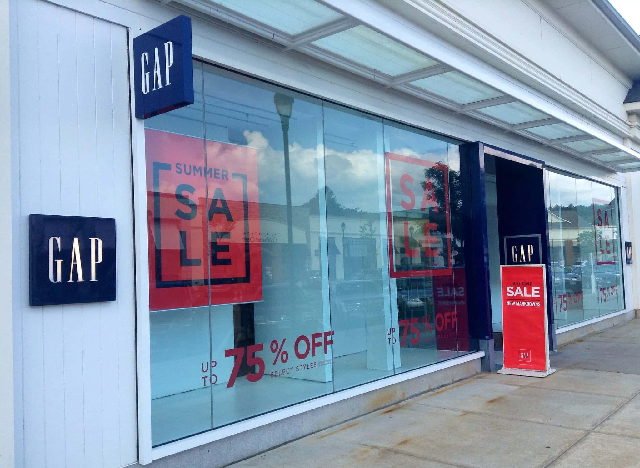間隙 桌布 壁紙 — Gap 店面正面，大型玻璃櫥窗展示醒目的紅色夏季促銷標誌，提供高達75%折扣，映襯著明亮的白天天空倒影。