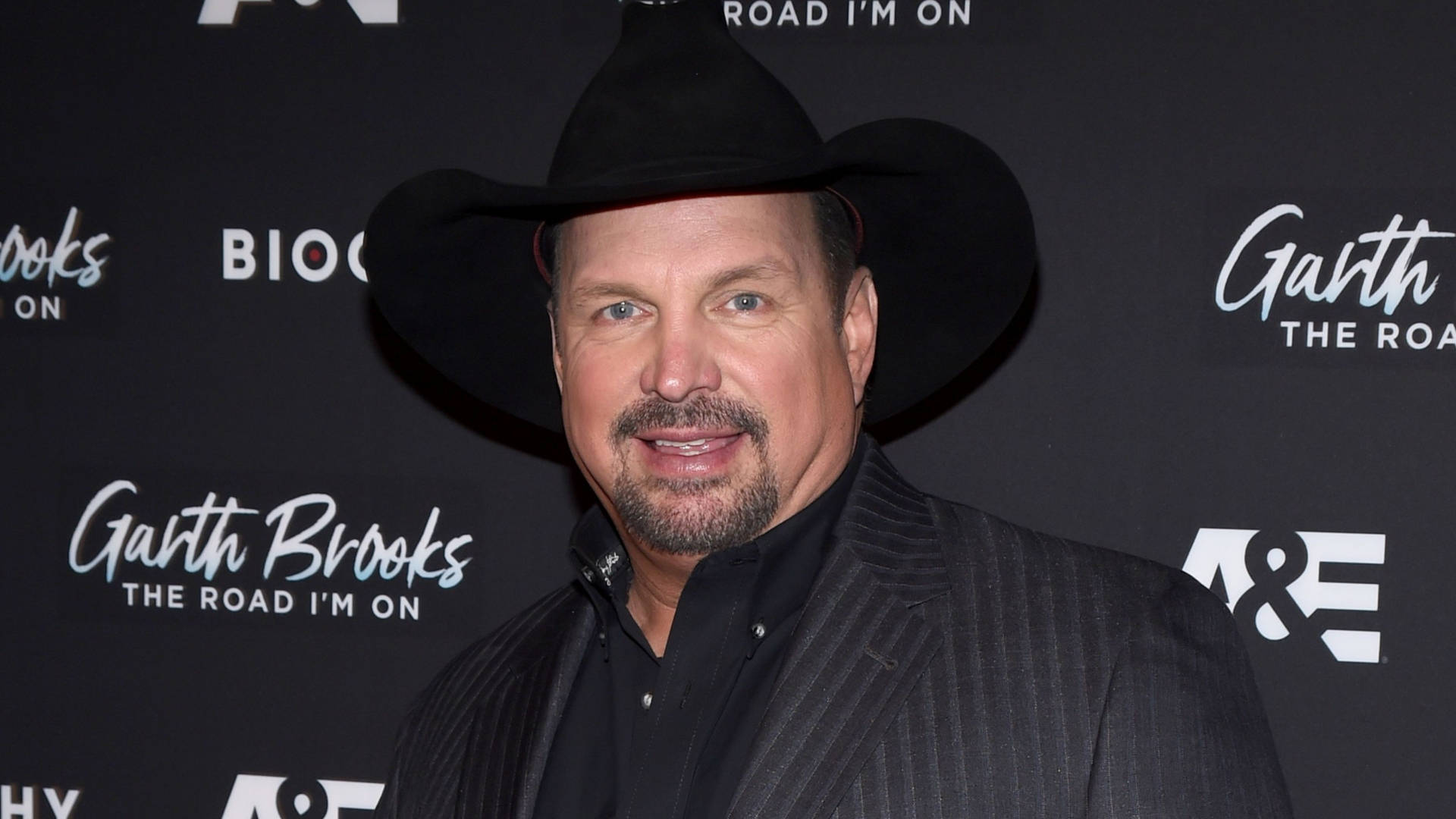 Singing 桌布 壁紙 — Garth Brooks 戴著黑色牛仔帽和細條紋西裝，在品牌活動背景前自信地微笑。