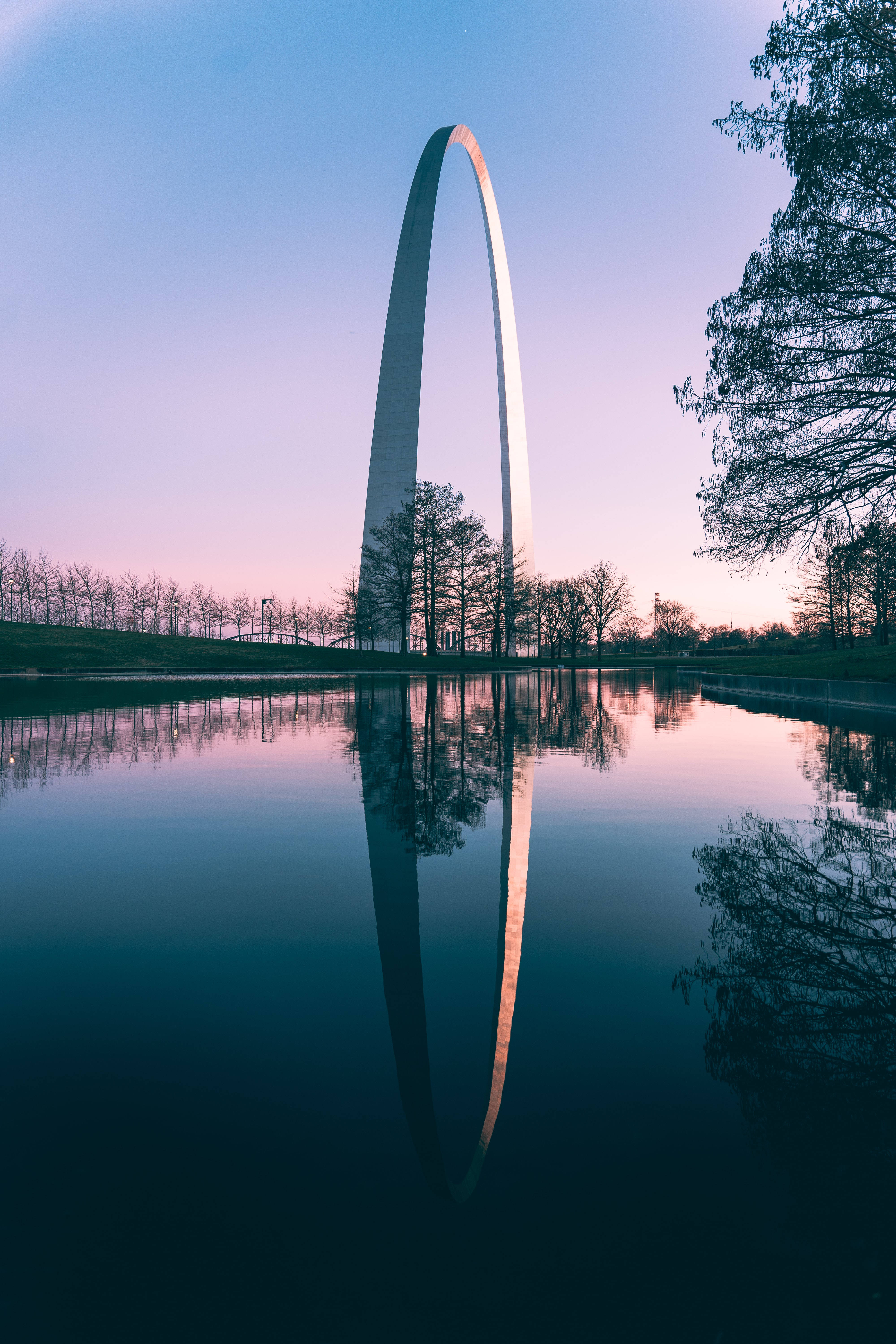建築 桌布 壁紙 — Gateway Arch 在暮色天空下高聳入雲，其完美的倒影映照在平靜的水面上，周圍是光禿禿的樹木。