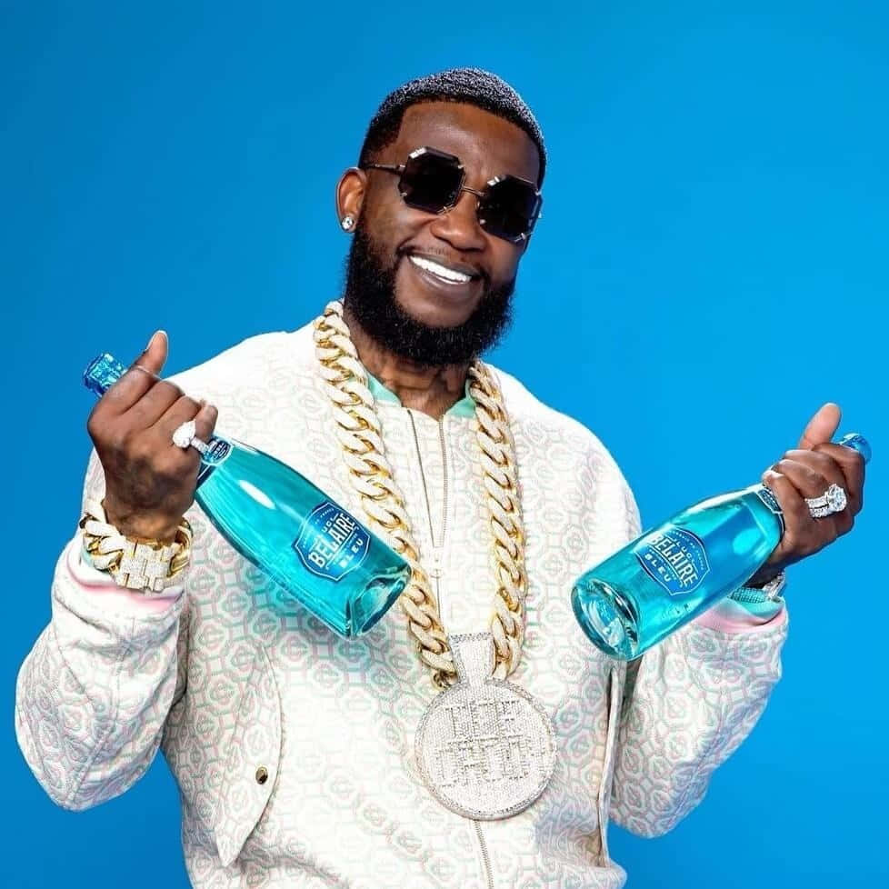  壁紙 — 一個戴著太陽眼鏡和金色鏈條的微笑男子，在純藍色背景前拿著兩瓶藍色Belaire酒。