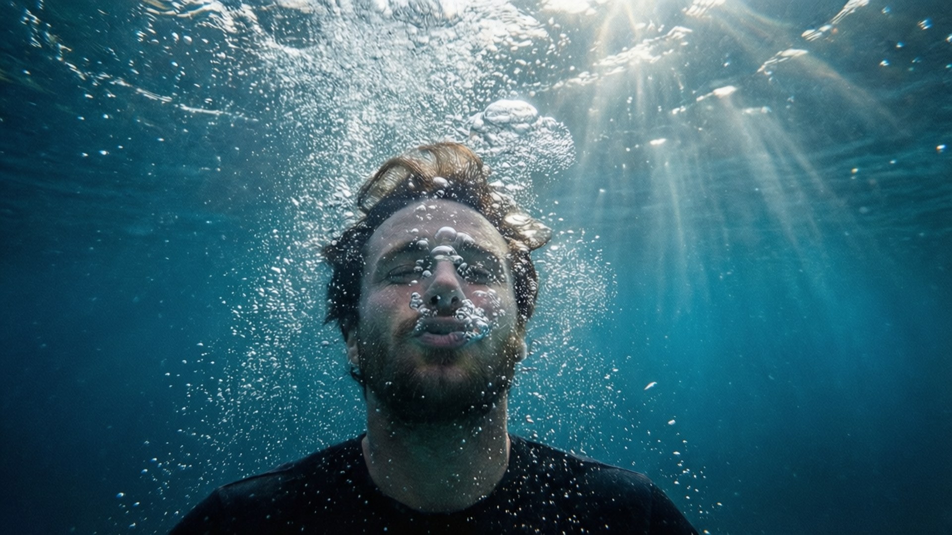 Underwater Breath 壁紙 — 一個留鬍鬚的男人在水底呼吸，陽光穿過他周圍的藍色海洋照射下來。