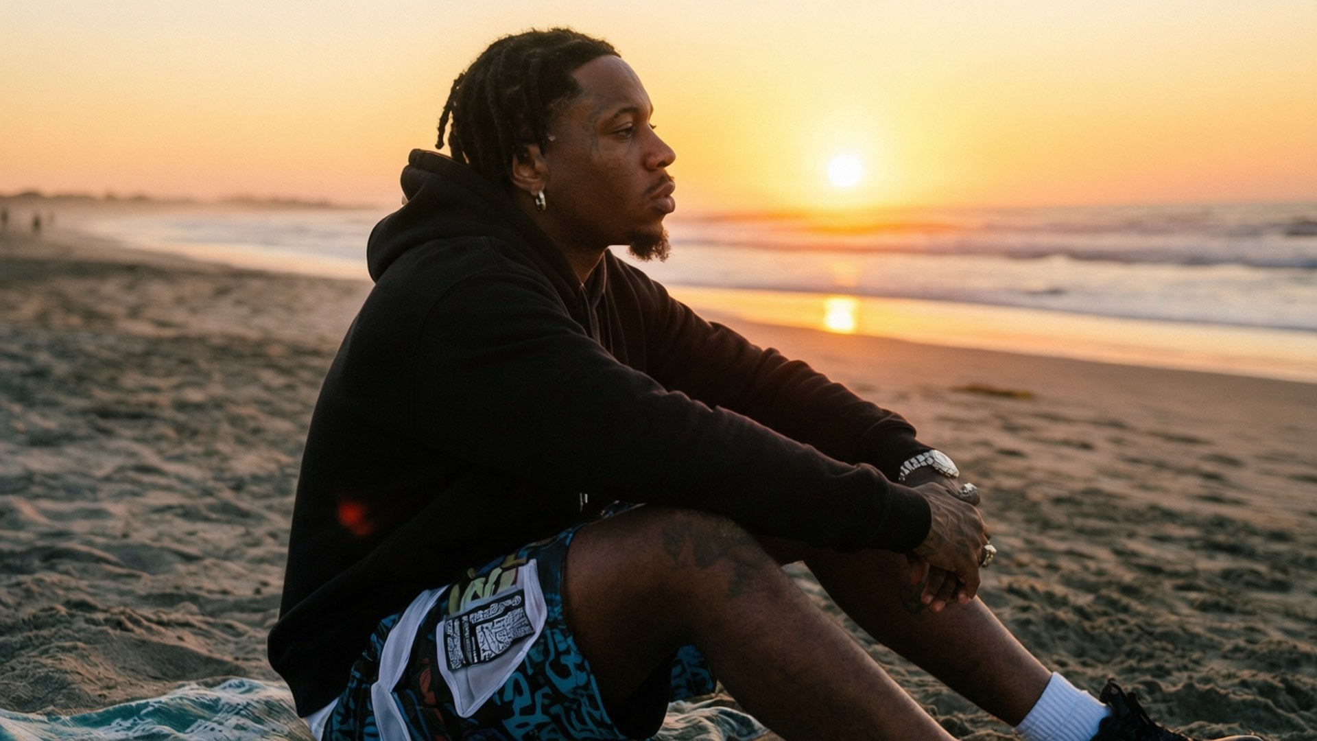 Beach Sunset 壁紙 — 一個留著辮子頭的男人坐在日落時分的沙灘上，穿著黑色連帽衫和圖案短褲，若有所思地凝視著大海。