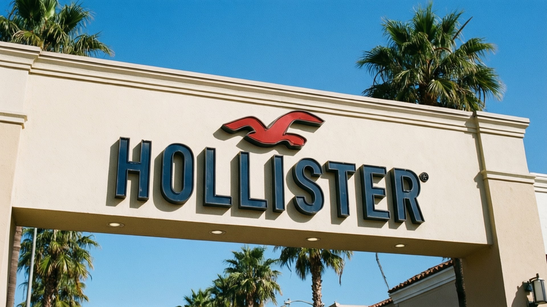 Store Sign California 壁紙 — 一個米色Hollister商店招牌，帶有紅色飛鳥標誌和藍色字體，矗立在晴朗藍天和棕櫚樹下。
