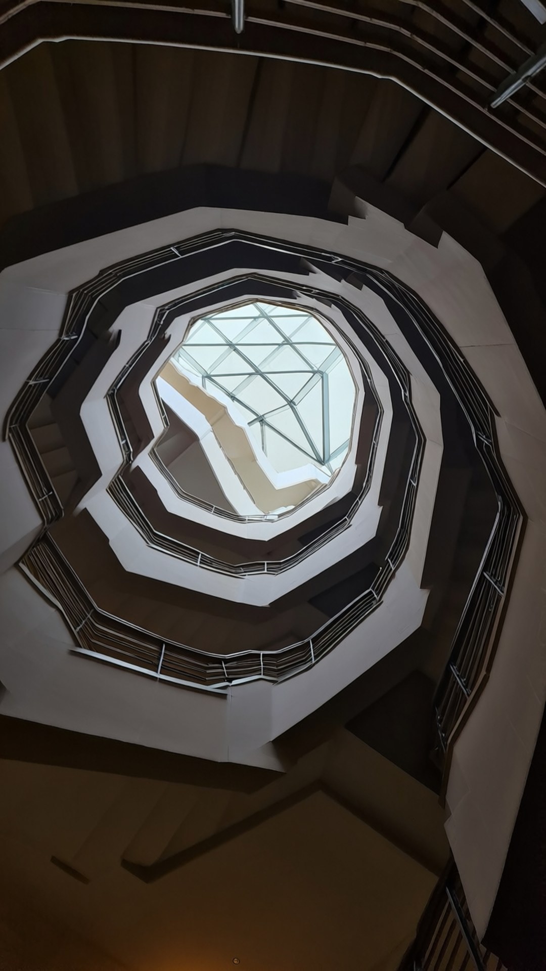 Al Hamra Tower Interior 壁紙 — 幾何螺旋樓梯，白色和深棕色階梯向上通往明亮的八角形天窗，配有玻璃板和金屬框架。