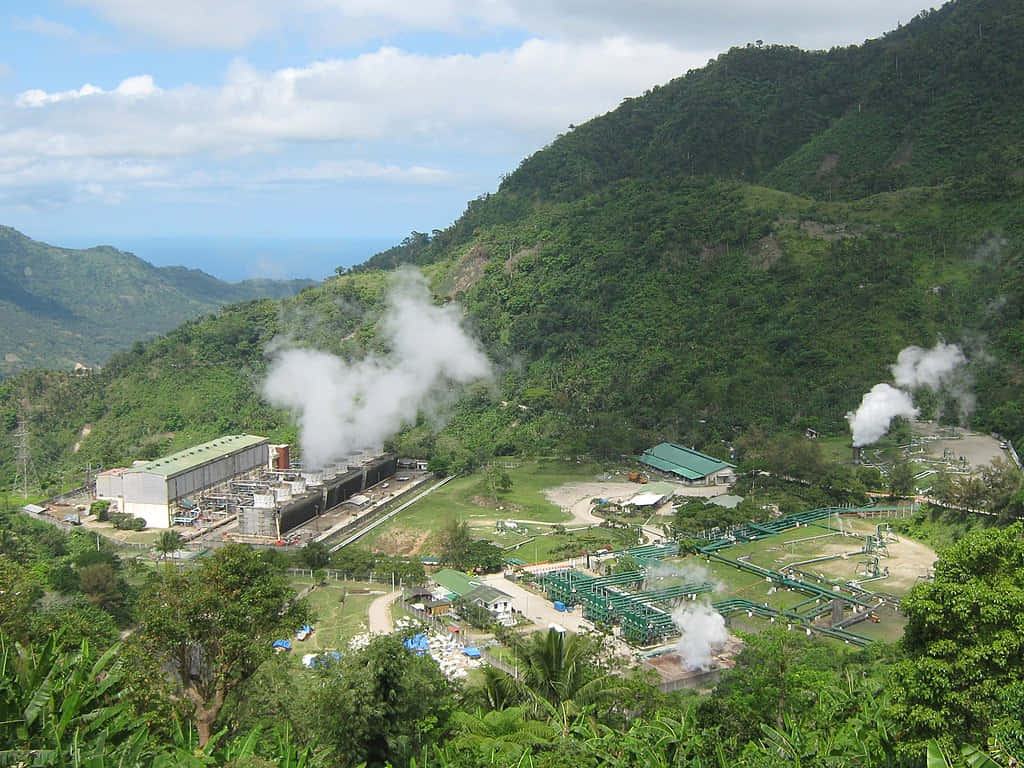  壁紙 — 一座地熱發電廠坐落在鬱鬱蔥蔥的綠色山脈中，在部分多雲的天空下，蒸汽從管道和建築物中升起。