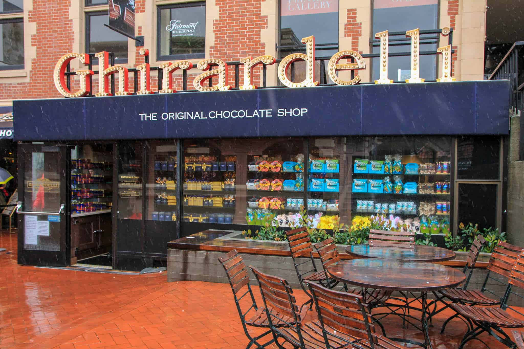 Chocolate 桌布 壁紙 — Ghirardelli 巧克力店在雨天，發光招牌和戶外座位在濕磚路上。