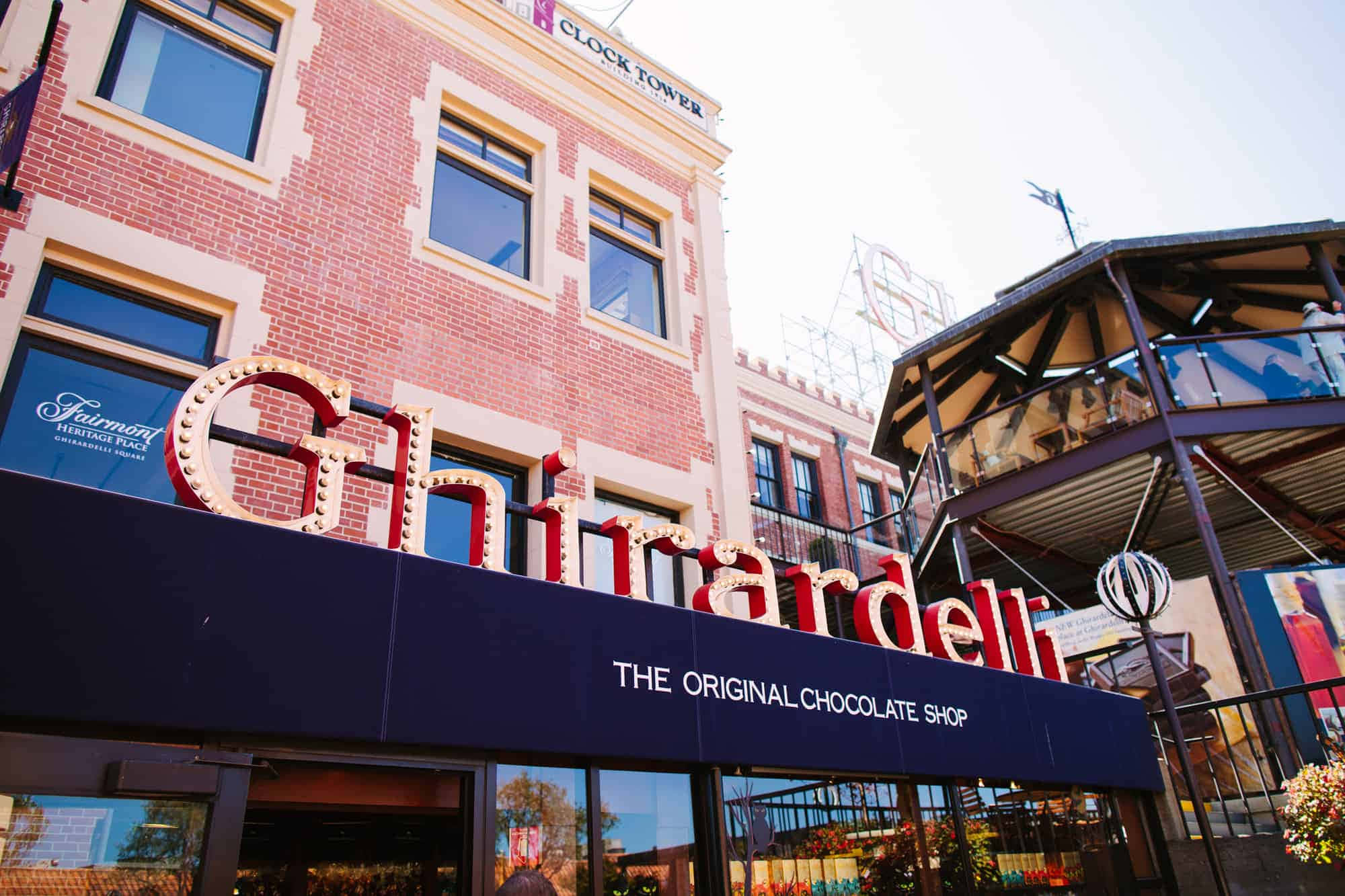 Chocolate 桌布 壁紙 — Ghirardelli 巧克力店的明亮紅色霓虹招牌點綴了一棟磚造建築，背景是晴朗天空、鐘樓和摩天輪。