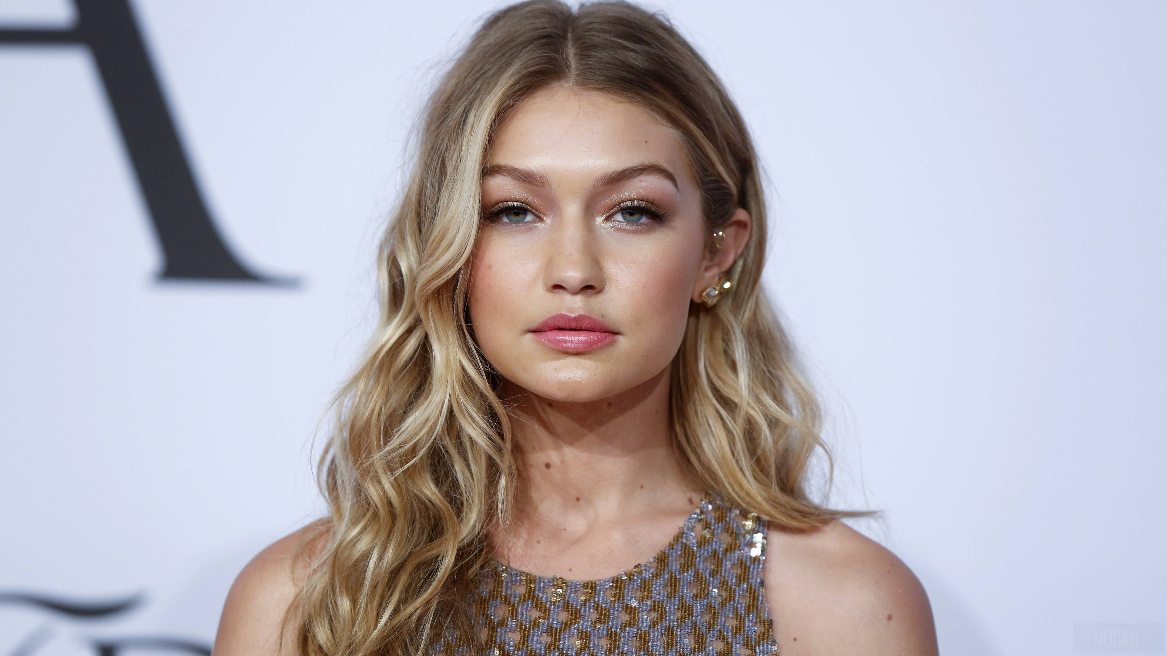 模特兒 桌布 壁紙 — Gigi Hadid 以波浪金髮和亮片上衣在白色背景前擺姿勢，背景上有黑色字樣。