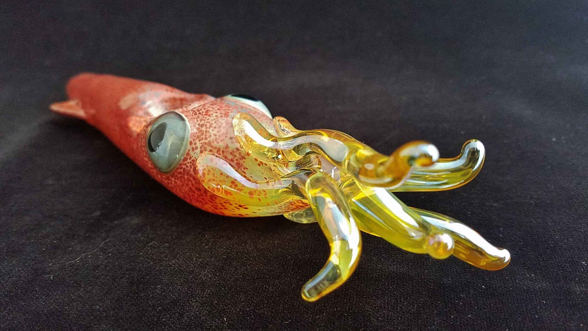  壁紙 — 一個充滿活力的玻璃魷魚雕塑，帶有紅色斑點的身體和半透明的黃色觸手，放置在深色布料表面上。