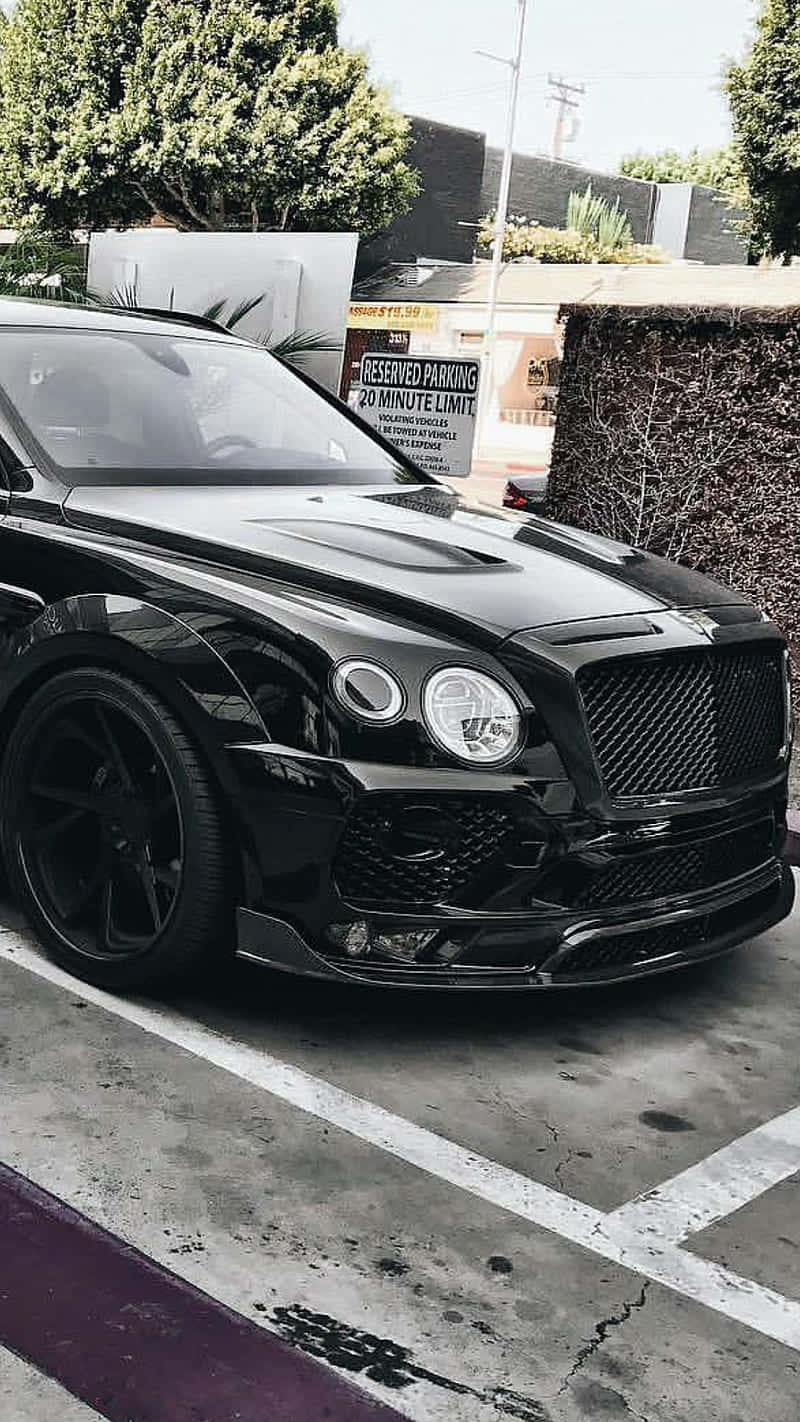 Bentley Bentayga 桌布 壁紙 — 一輛亮黑色賓利Bentayga停放在陽光普照的城市空地，背景有樹木和預留停車標誌。