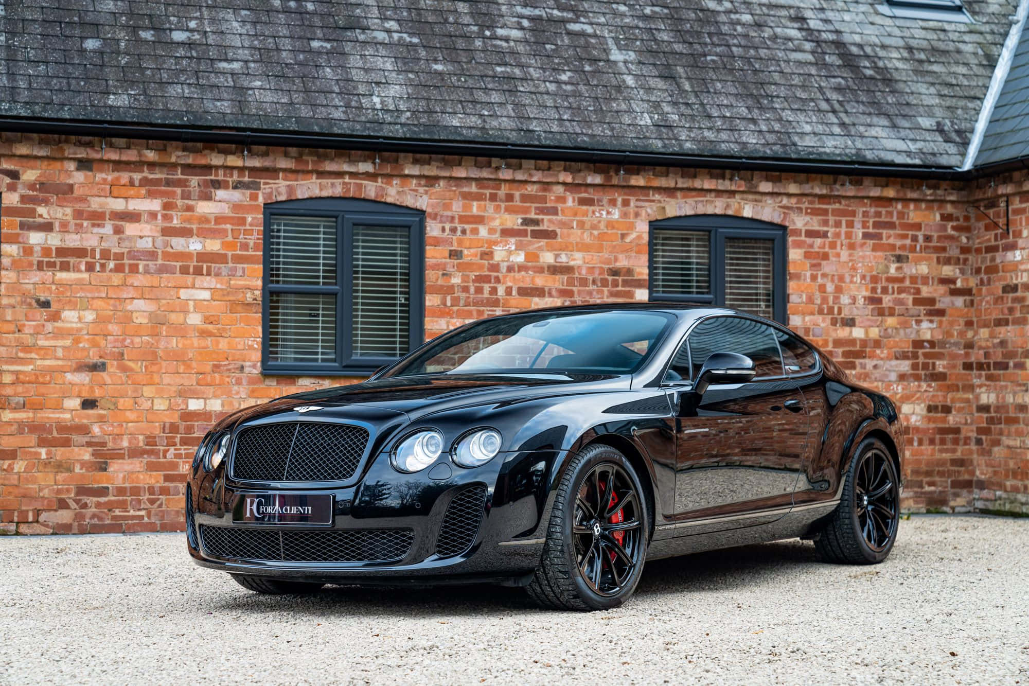 Bentley Continental Supersports 桌布 壁紙 — 一輛亮黑色賓利 Continental GT 雙門跑車停放在紅磚建築前的碎石地上，建築物有深色窗戶。