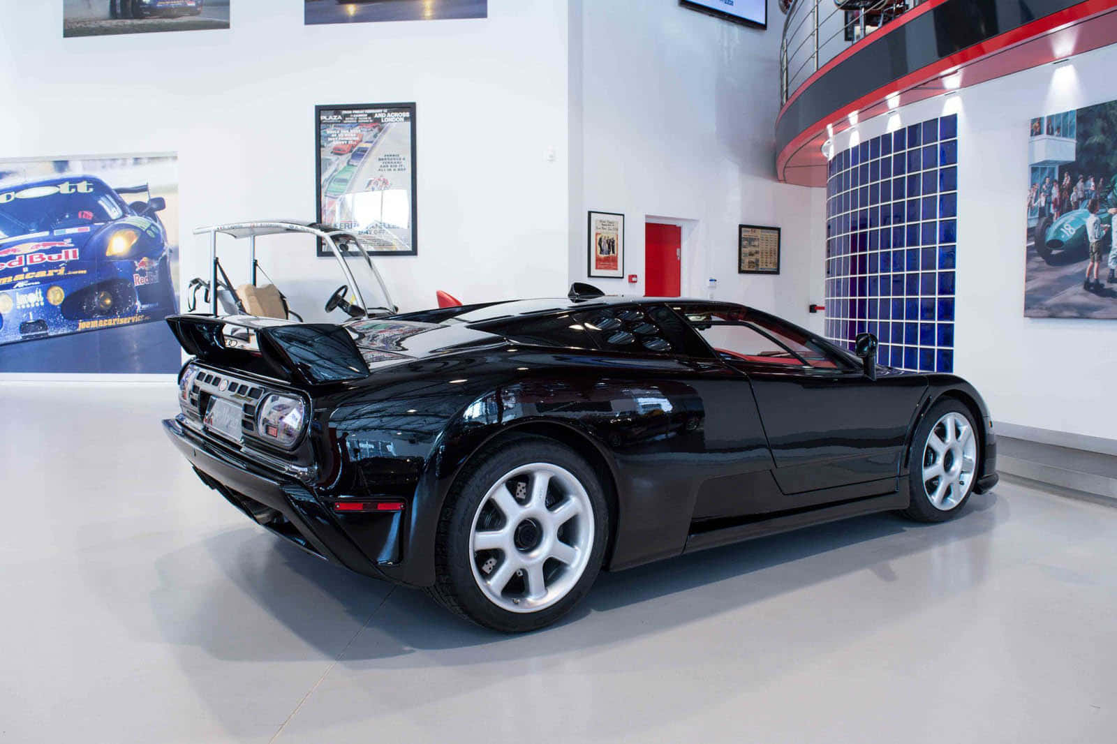 Bugatti Eb110 桌布 壁紙 — 一輛亮黑色Bugatti EB110超級跑車在明亮現代汽車博物館中展出，地板反射光線，牆上掛有賽車海報。
