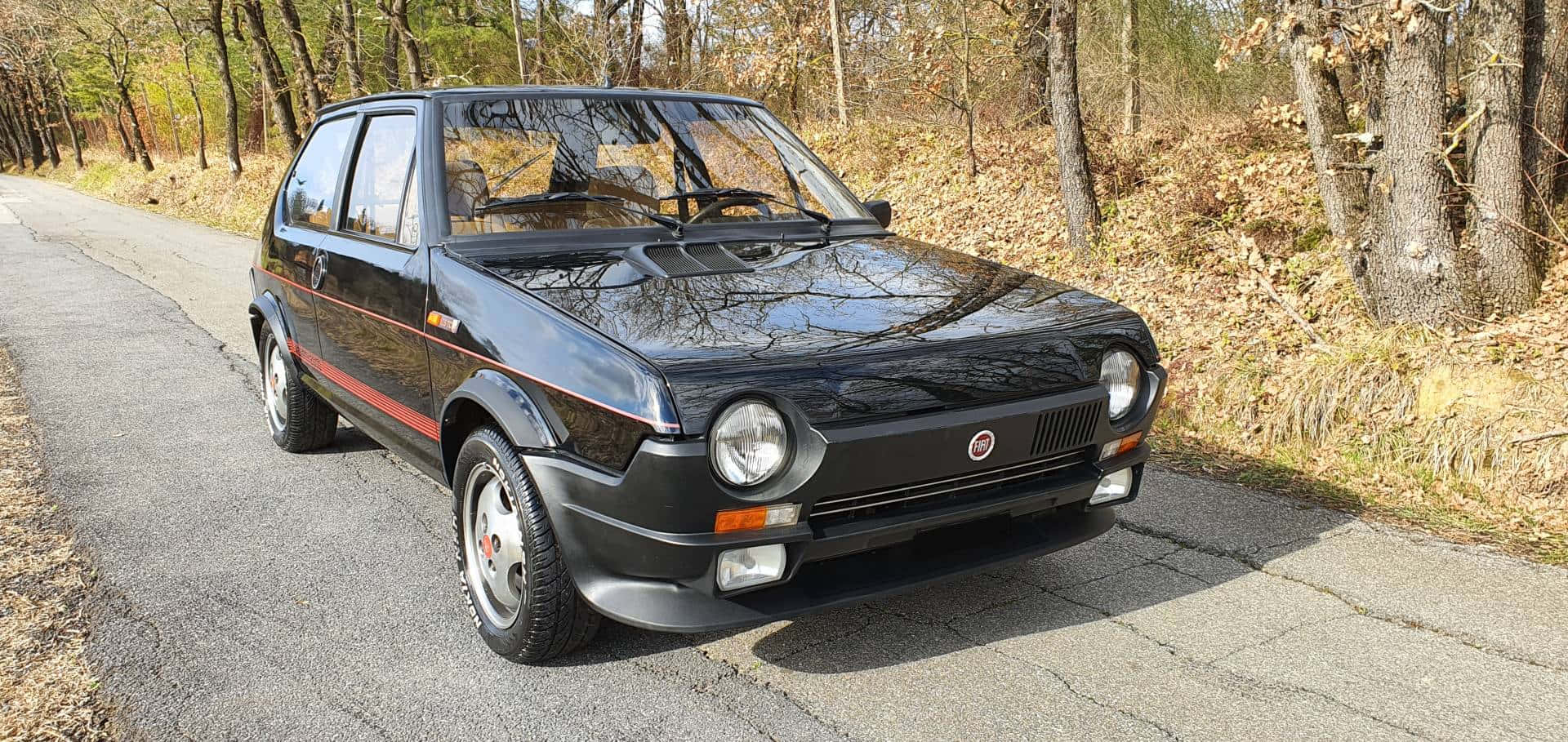 Fiat Ritmo 桌布 壁紙 — 一輛亮黑色的飛雅特 Uno Turbo 停放在一條裂開的柏油路上，兩旁是秋天的禿樹。