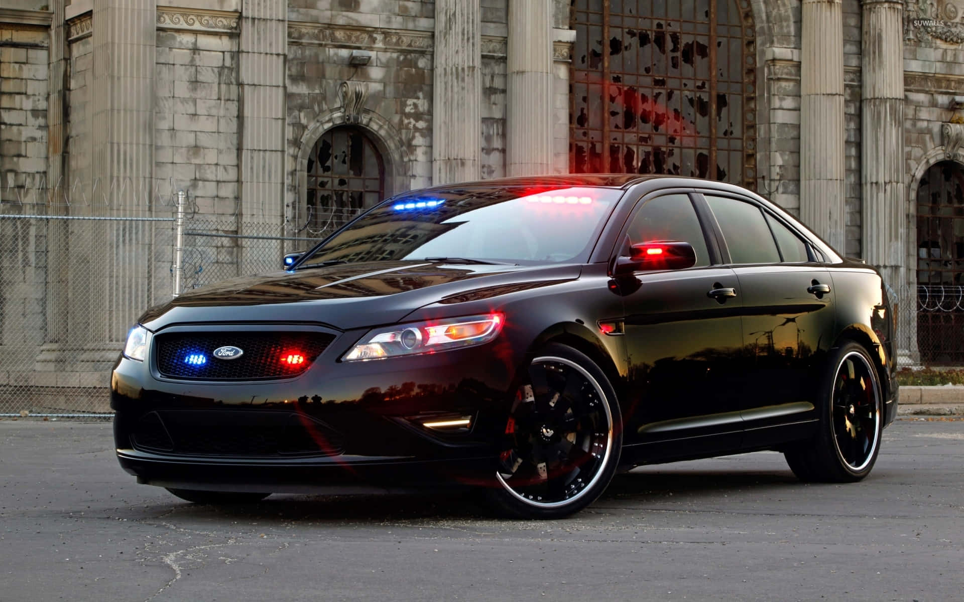 Ford Taurus 桌布 壁紙 — 一輛亮黑色福特警車，閃爍著紅藍警燈，停在一座風化的石建築前。