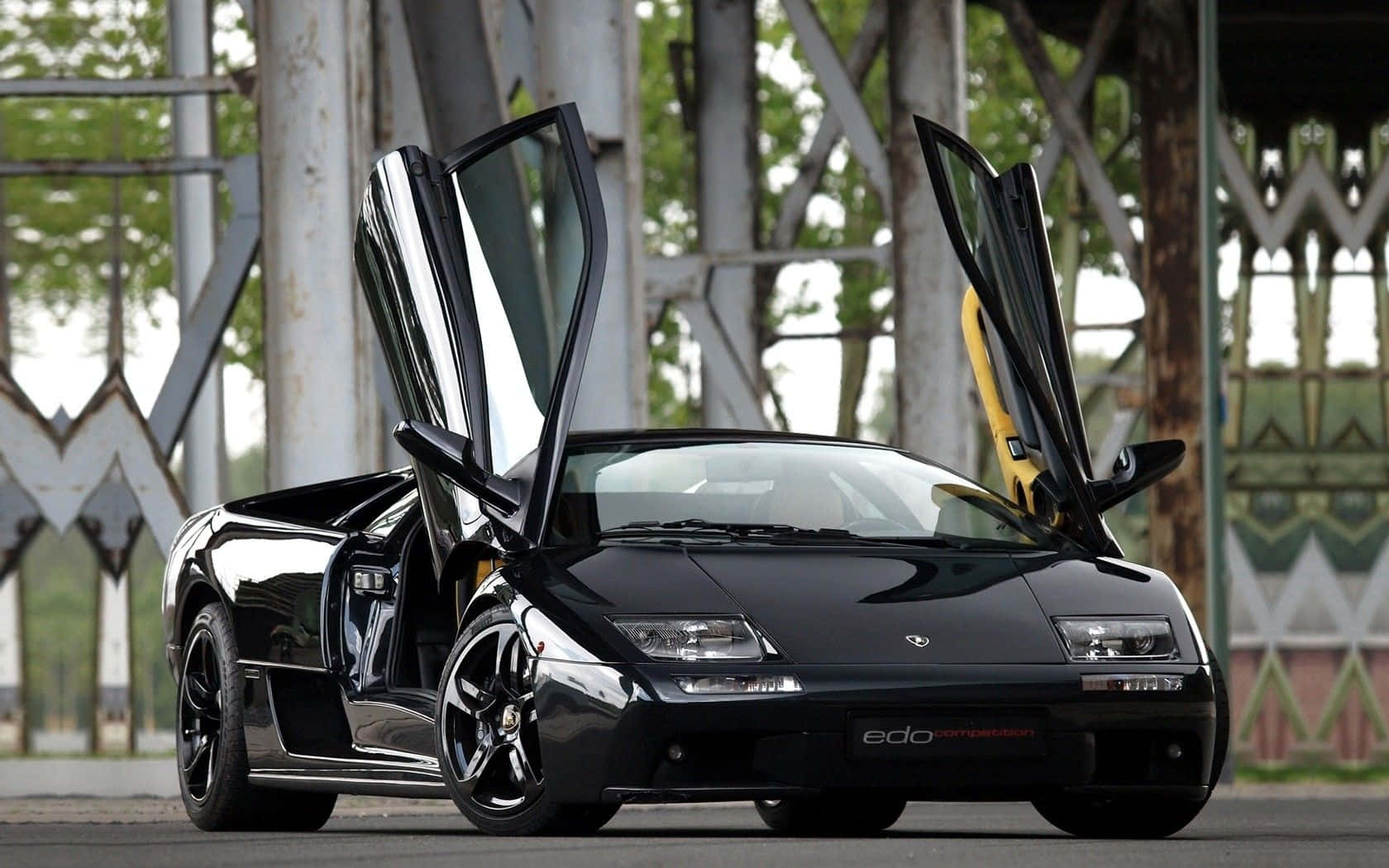 Lamborghini Diablo 桌布 壁紙 — 一輛亮黑色藍寶堅尼 Diablo 打開鷗翼車門，停在金屬橋下方，背景有綠色植物。
